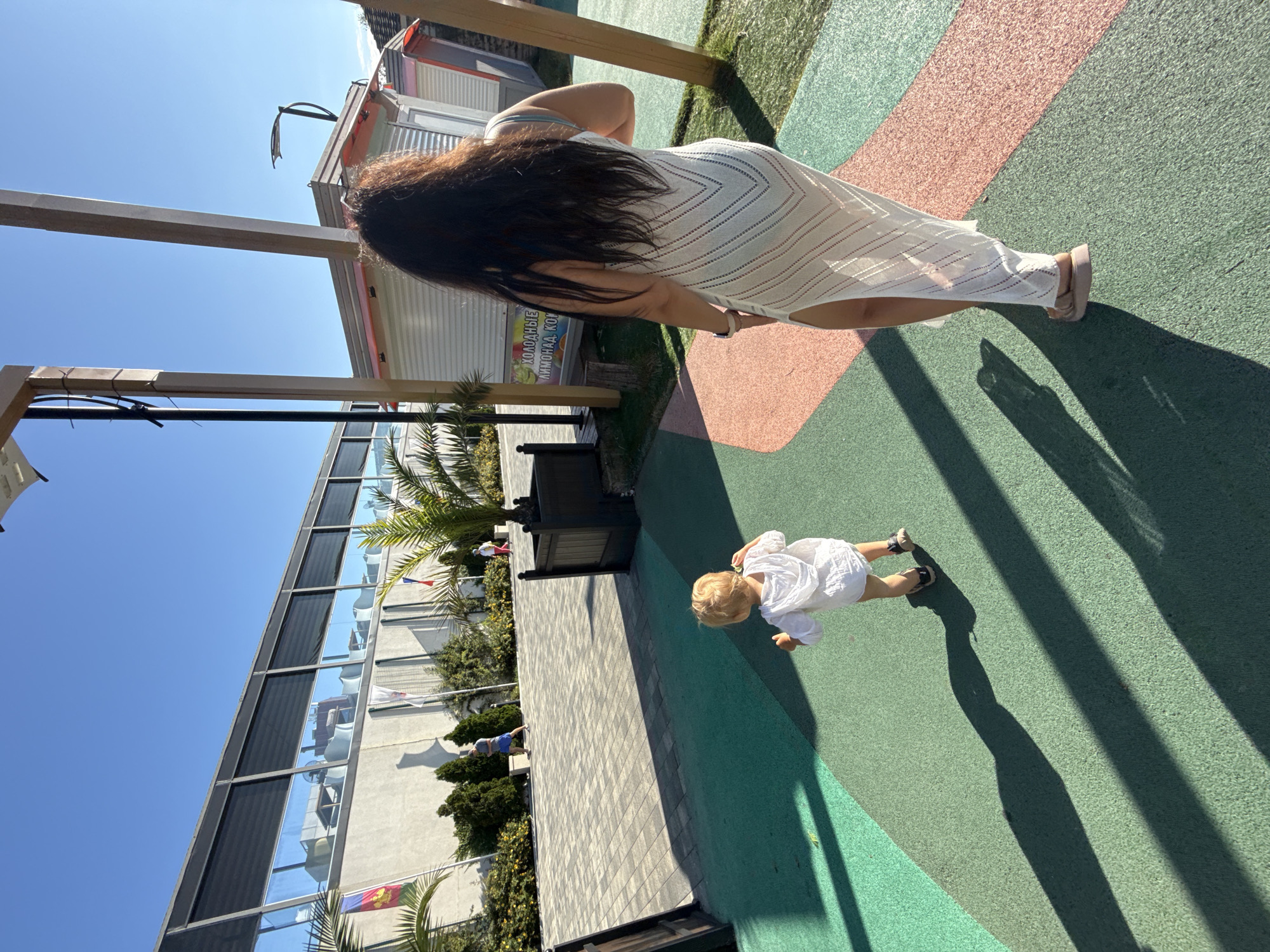 The image depicts an outdoor scene, likely a promenade or public area, with a focus on two individuals, an adult and a young child, walking away from the camera. The ground surface is paved with sections of green and pink material. In the background, there is a modern building with large windows reflecting the clear blue sky. Adjacent to this building are palm trees and other greenery. To the right, a small, white kiosk-like structure with pink trim is visible, along with wooden pillars supporting overhead elements. The lighting suggests it is a sunny day, casting distinct shadows on the ground. The presence of flags and the architectural style of the kiosk suggest a resort or leisure area, consistent with locations found in Sochi, Russia.