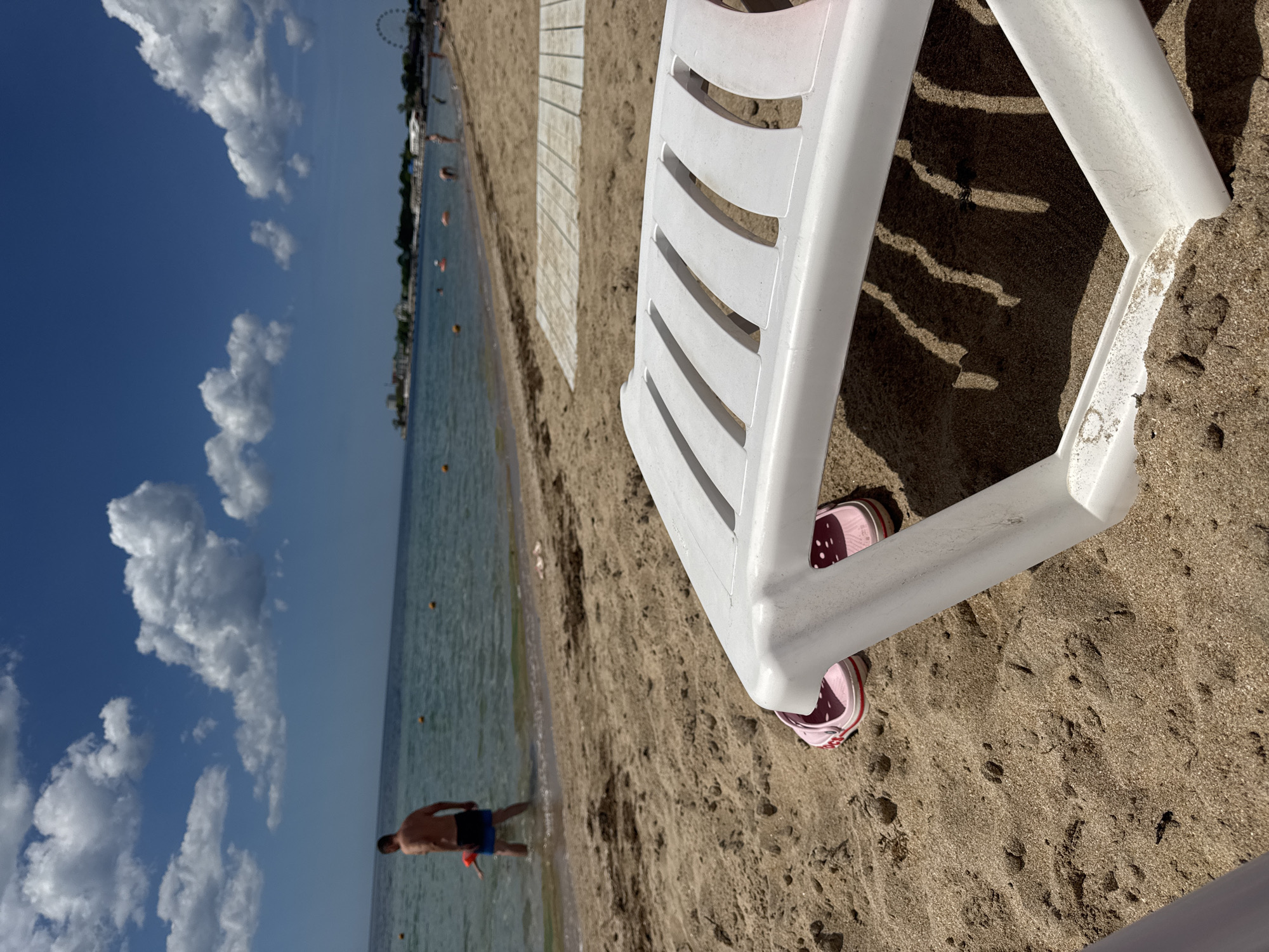 The image depicts a beach scene in Yevpatoriya, Ukraine. A white plastic beach chair sits on the sand, with a pair of pink shoes tucked beneath it. In the background, a man is wading in the water. Buoys are visible in the water, and a boardwalk runs along the beach. The sky is blue with scattered clouds.