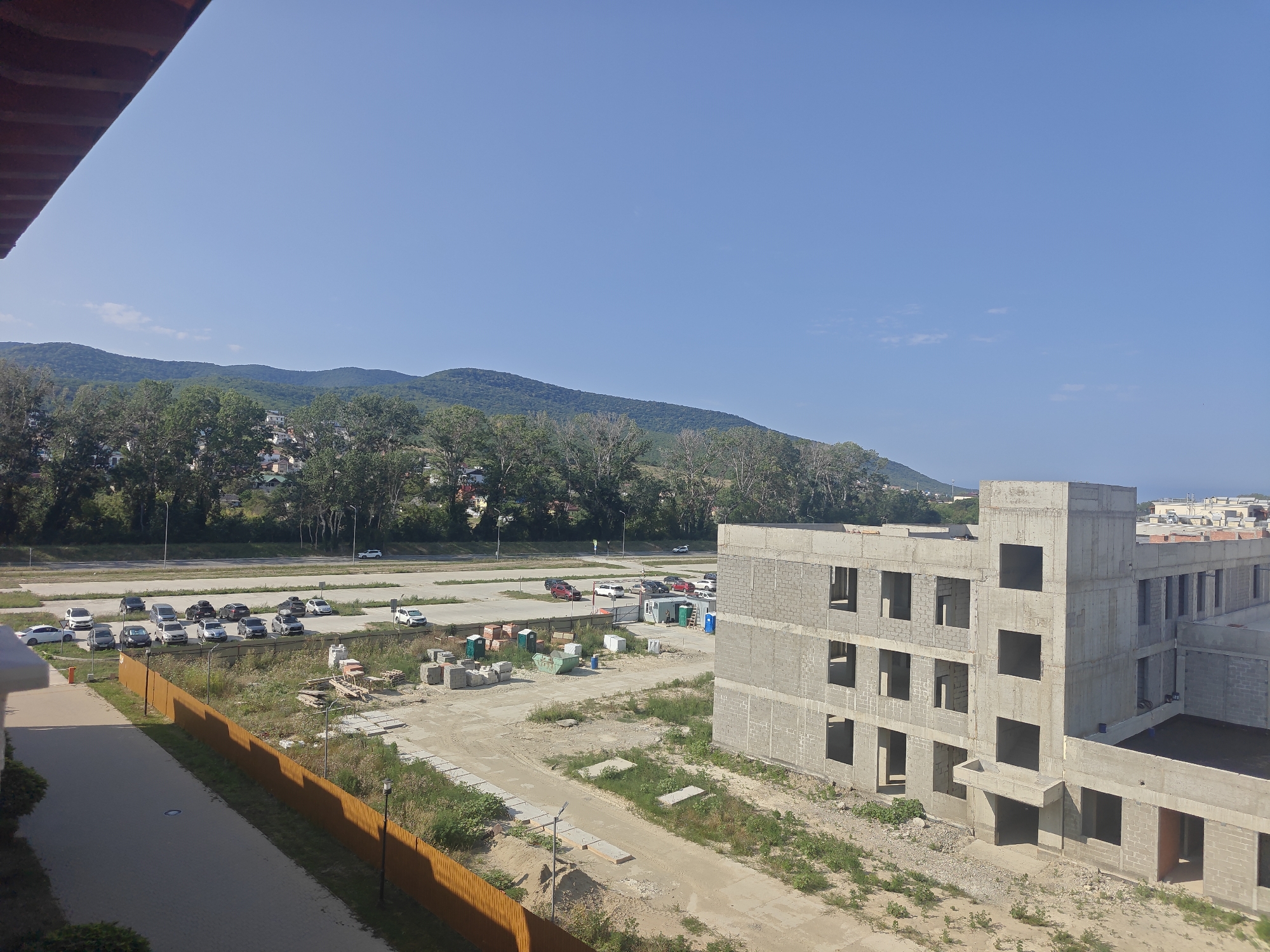 In the foreground, a section of a reddish-brown roof overhang is visible on the left. Beyond this, a paved walkway runs diagonally from the lower left towards the center right. This walkway is bordered by a low, orange wooden fence on its right side.
The midground is dominated by a construction site. A large, unfinished concrete building with multiple stories and empty window openings is a prominent feature. Various construction materials, including stacks of bricks, concrete blocks, portable toilets, and construction trailers, are scattered around the site. Several vehicles are parked in a lot to the left of the construction area, including a white car, several gray cars, and a few dark-colored vehicles. A road runs along the far edge of this parking area.
The background depicts a densely wooded area with numerous green trees, behind which are hills and mountains covered in vegetation. A few buildings are visible nestled among the trees on the hillsides. The sky above is clear and blue with a few wispy white clouds. The overall scene suggests a suburban or semi-rural environment undergoing development. The image was taken on a sunny day.