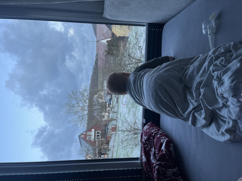 A child in a sleep sack looks out a window at a snowy landscape. Outside, a red house with a white facade and bare trees are visible. The sky is overcast with heavy, dark clouds. A bottle with a tube attachment rests on the bed next to the child. A patterned blanket is draped on the bed. The photo is taken from a slightly elevated position.
