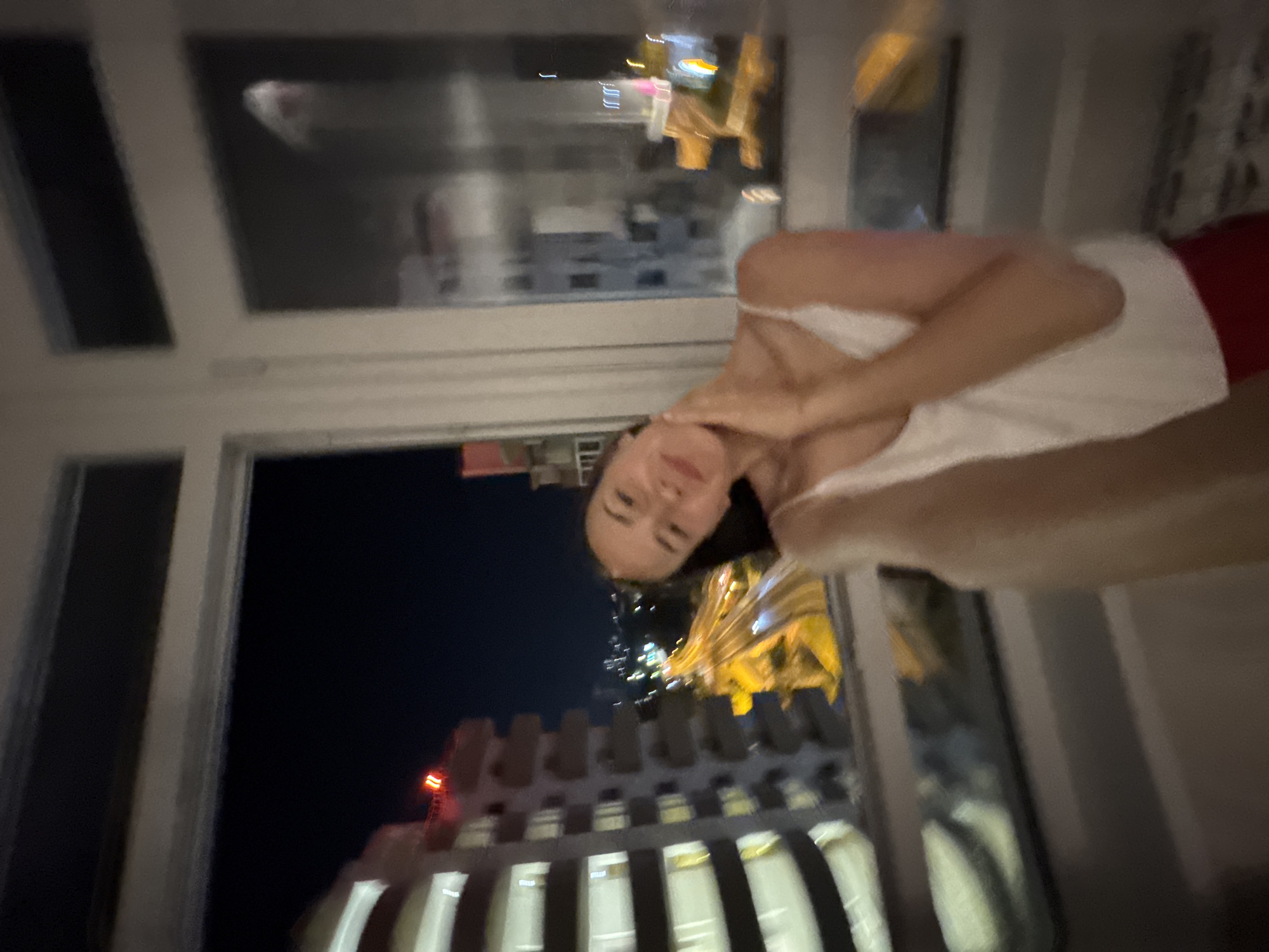 A woman is captured from the chest up, posing for a selfie in an interior space with a large window overlooking a city at night. She is wearing a white tank top and red pants. The window reveals a view of illuminated buildings, a highway with car lights, and a dark night sky. The scene is slightly blurred, suggesting movement or a shallow depth of field. The architecture visible outside suggests an urban environment, consistent with Sochi, Russia.