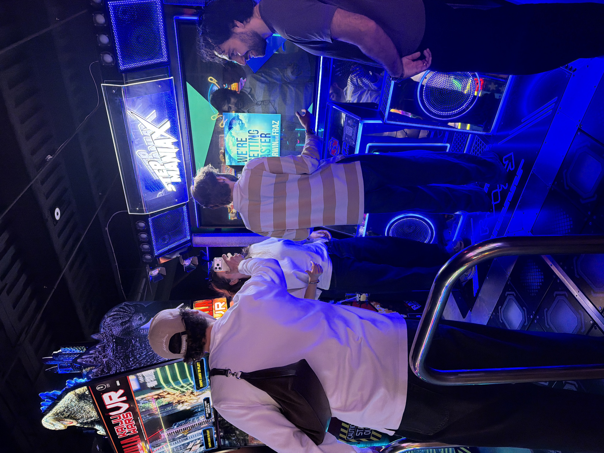 The image depicts four individuals, three males and one female, engaged with arcade games in a dimly lit, neon-blue illuminated establishment. Two individuals are standing in front of a "KAIJU WARS VR" game, while two others are positioned before a "STEP MANIAX" game. One person is actively capturing a photo or video with a smartphone. The games feature large screens displaying animated content and colorful graphics. The flooring of the "STEP MANIAX" area consists of illuminated pressure-sensitive pads arranged in a grid pattern, typical of dance or rhythm games. The environment suggests an arcade or entertainment center. The presence of "Montreal, Canada" in the prompt suggests this arcade is located in that city.