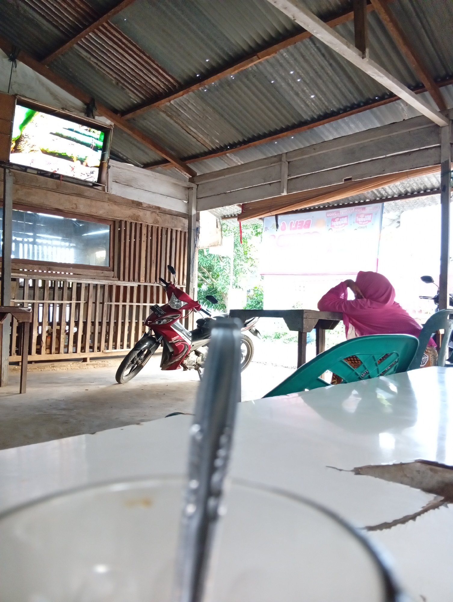 The image depicts an outdoor covered seating area, possibly a warung or small eatery, in Lhokseumawe, Indonesia. A red and black motorcycle is parked to the left, facing the viewer. In the background, a person wearing a pink hijab is seated at a table, their head resting on their arms. Behind them, a banner with text is visible. The structure has a corrugated metal roof supported by wooden beams. A television screen is mounted on the wall, displaying an image. The foreground shows a white table with a blurred object that appears to be a utensil. The ground is concrete.