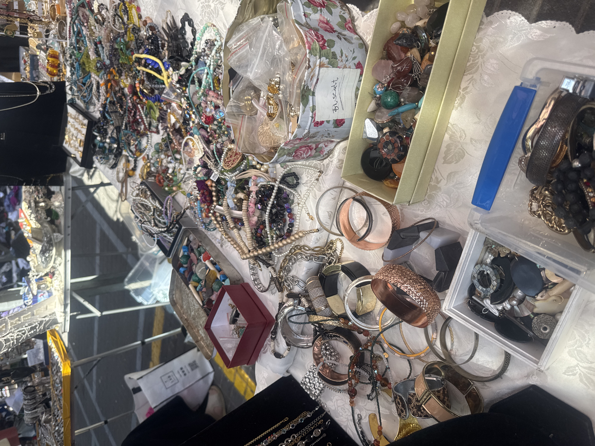 The image shows a table laden with various pieces of jewelry, including necklaces, bracelets, and earrings. These items are displayed in boxes and on the table's surface, which is covered with a white patterned cloth. A floral-patterned tin is visible, labeled "Biscuit." The location is likely a market or fair, possibly in Tel-Aviv, Israel, indicated by the type of jewelry and market-like setting. The image suggests a commercial activity, perhaps a sale of vintage or handmade jewelry. The scene is set outdoors, as inferred from the lighting.