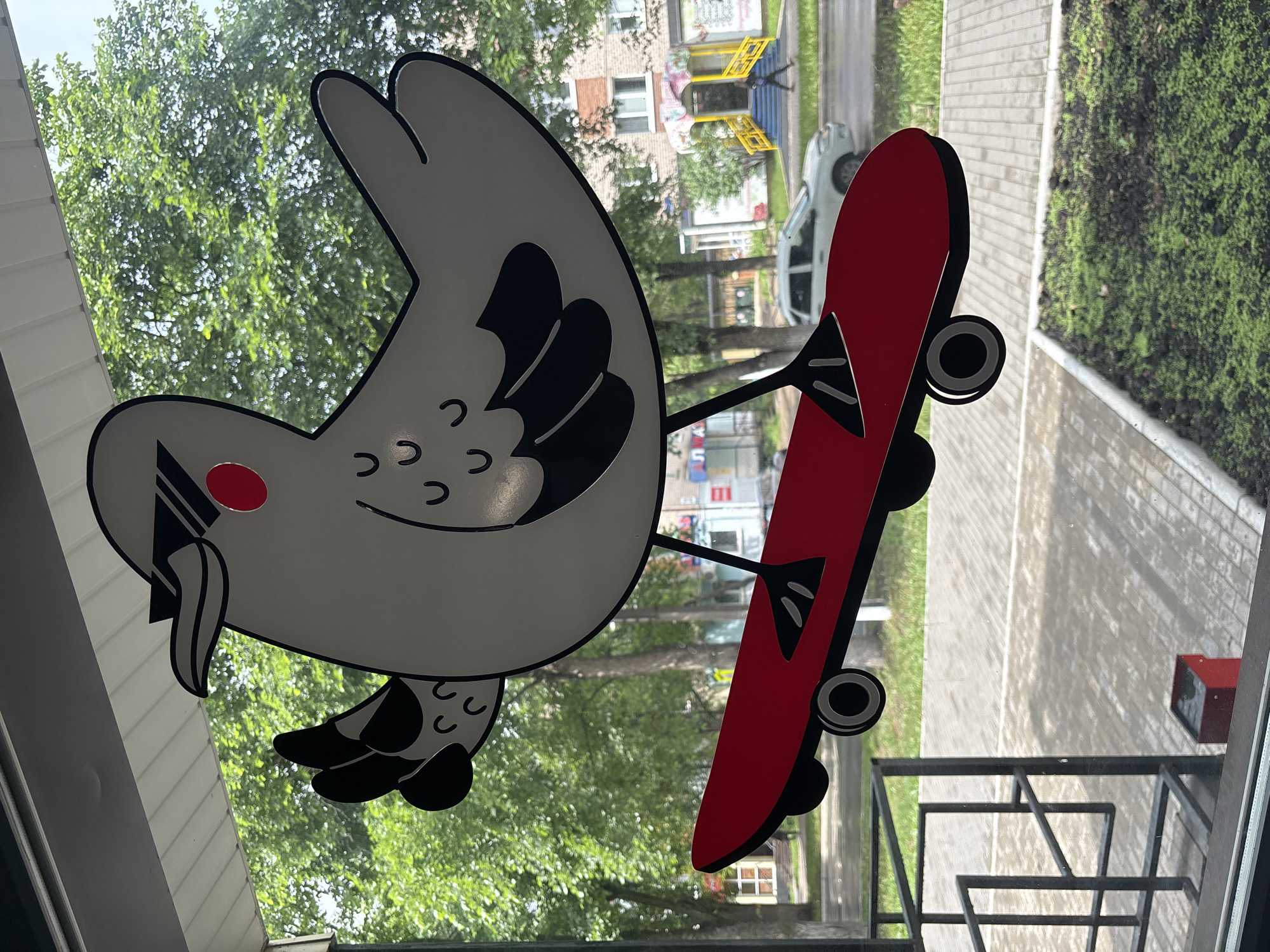 A decorative sticker of a cartoon bird wearing sunglasses and riding a red skateboard is affixed to a window. The bird is predominantly white with black accents on its wings, beak, and eyes, and a red circle for a cheek. It is depicted in a dynamic pose, with one wing raised as if waving. The skateboard is red with black wheels and accents.
Through the window, a street scene in Chaikovsky, Russia is visible. Lush green trees line the sidewalk, and a light-colored car is parked on the street. In the background, there is a building with a yellow entrance and a multi-colored awning. The ground outside the window consists of a paved sidewalk and a patch of green grass. The overall impression is that of a whimsical decoration in a semi-urban setting.