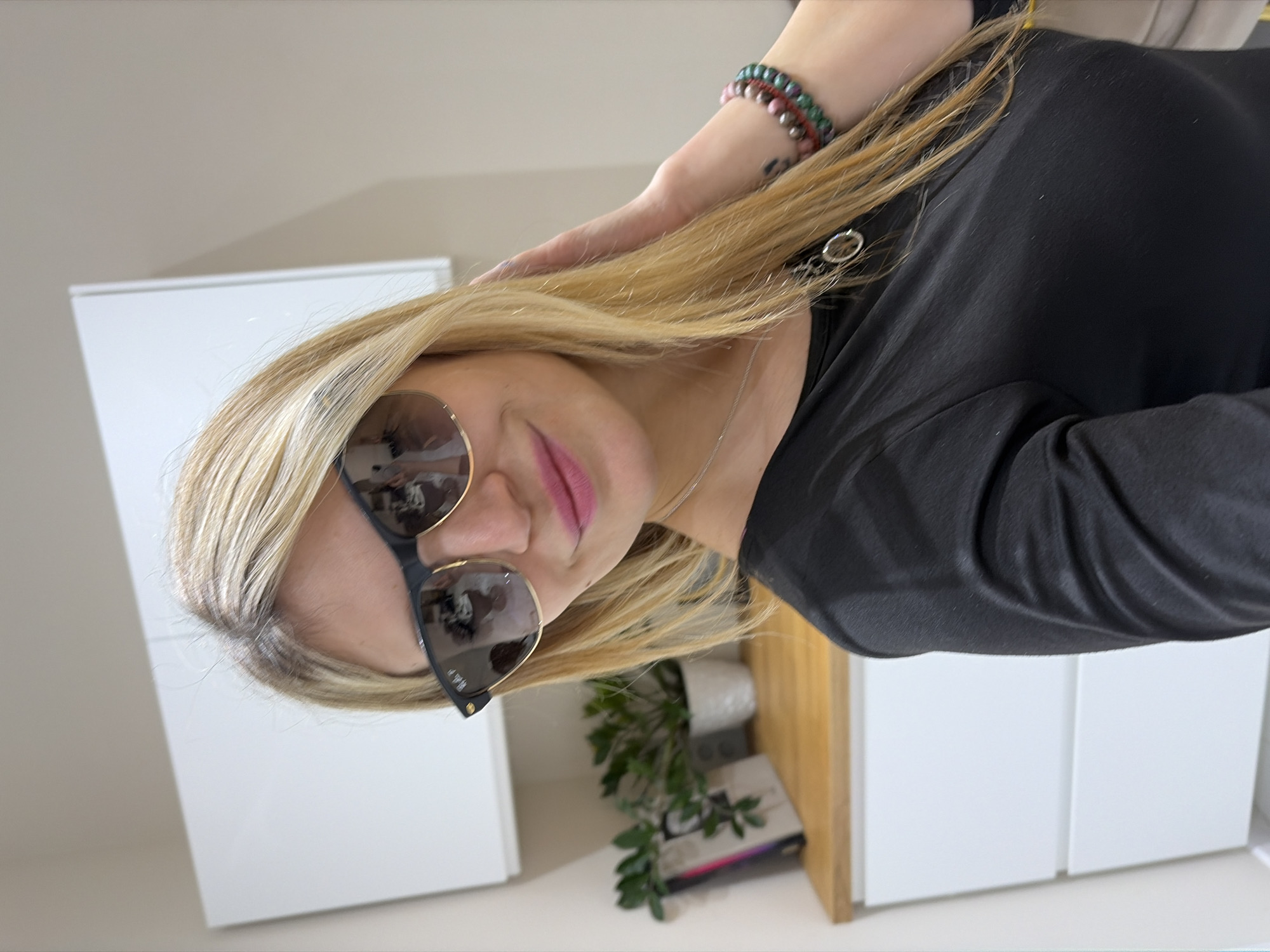 A woman with blond hair and sunglasses is shown indoors. She is wearing a black shirt and has a bracelet on her wrist. There are white cabinets in the background, along with a small green plant in a white pot. There is a shelf with books. The location may be Budva, Montenegro.