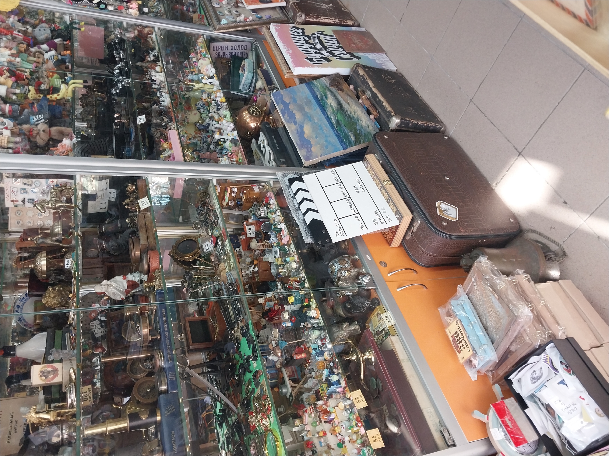 The image presents an overhead view of a cluttered display of various items, likely within a market stall, antique shop, or flea market. The scene is dominated by glass display cases, filled with a diverse collection of small objects.
The cases are stacked vertically. The upper cases predominantly feature numerous small figurines, including dolls, miniature animals, and various toy-like items. The middle and lower sections of the display cases hold a range of metallic and antique-style objects, such as ornate door handles, keys, small telescopes, compasses, miniature decorative statues, and what appear to be old coin or medal collections. Prices in various denominations are visible on small tags attached to some items.
On the counter space directly above the glass cases, there are several larger items. These include old books with ornate covers, one featuring the Russian title "НАЦИОНАЛЬНЫЙ ВОПРОС" (National Question) and another "ПРАКТИЧЕСКОЕ КОРАБЛЕВОЖДЕНИЕ" (Practical Ship Navigation). A painting depicting a seascape, a film clapperboard with English and Chinese text ("PRODUCTION," "DIRECTOR," "CAMERAMAN," "片名" (film title)), and two vintage-style leather suitcases are also present. On the right side of the counter, various small packaged items, some with prices like "380" and "85," are displayed next to a small decorative ship's wheel.
A prominent sign in Russian reads "БЕРЕГИ ХОЛОД - ЗАКРЫВАЙ ДВЕРЬ" (Protect from cold - Close the door), suggesting the need to maintain temperature control. The flooring is made of light-colored tiles. The overall impression is that of a rich and eclectic collection of goods, likely intended for sale.