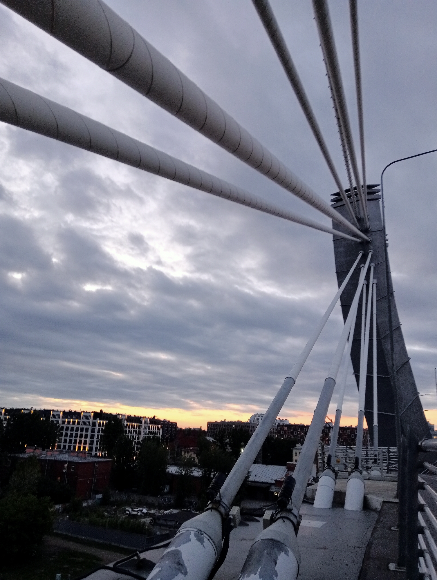 The image shows a view from the top of a cable-stayed bridge in Saint Petersburg, Russia, looking towards the city skyline at dusk. The foreground is dominated by the bridge's massive concrete pylons and the thick, coiled cables that suspend the bridge deck. These cables are white with a gray spiral pattern. The pylons are dark gray and angular.
In the middle ground, a portion of the city is visible, characterized by multi-story buildings with windows reflecting the fading light of the sunset. Some trees and darker, lower-rise structures are interspersed among the buildings. The sky is cloudy, with hints of orange and yellow peeking through the gray, indicating the sun is low on the horizon. The overall atmosphere is somber due to the overcast sky and the time of day. No people or vehicles are visible in the image. The infrastructure suggests a modern, urban environment.