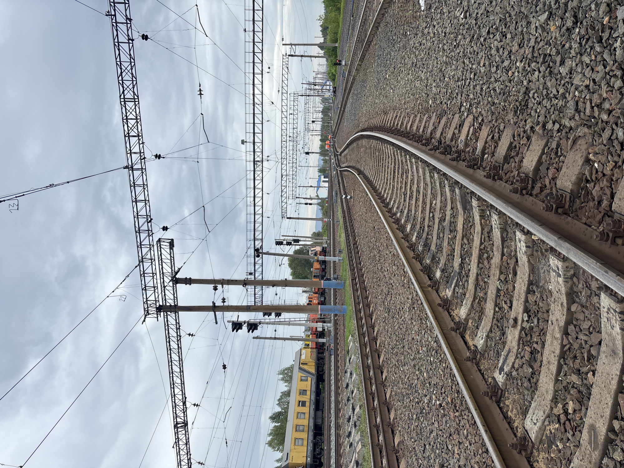 The image captures a railway yard with multiple tracks and overhead catenary systems in Borodino, Russia. In the background, several trains are visible, including a yellow passenger train and several orange locomotives. The tracks are laid on ballast, with concrete sleepers supporting the rails. Overhead power lines are strung between poles, suggesting an electrified railway line. The sky is overcast with clouds. The scene appears to be operational, with trains present and infrastructure in place for railway traffic. The environment is typical of a railway yard, characterized by tracks, trains, and electrical infrastructure.