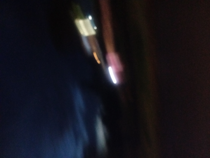 Blurry streaks of light streak across a dark background. A thicker, mostly obscured shape appears at the top, exhibiting white and green. A thinner, white line appears lower down, creating a sense of movement. Shades of blue, brown, and black dominate the indistinct scene, yielding an abstract effect.