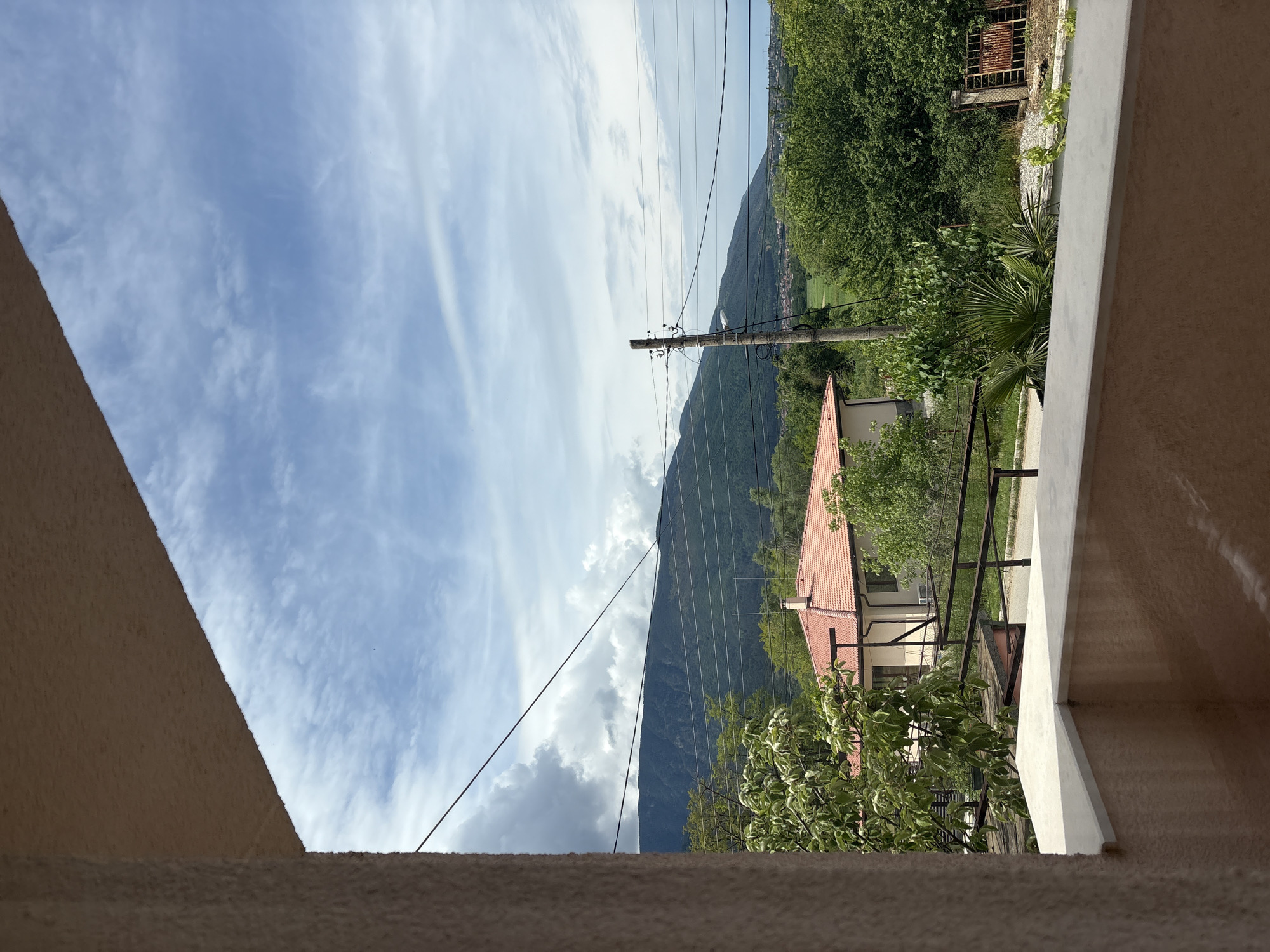 The image shows a view from a balcony in Saparevo, Bulgaria. In the distance, a mountain is visible, partially obscured by clouds and traversed by power lines connected to a utility pole. A house with a red tile roof is situated amongst trees on the hillside. The balcony is constructed of stucco and has a railing. The sky is blue with scattered white clouds.