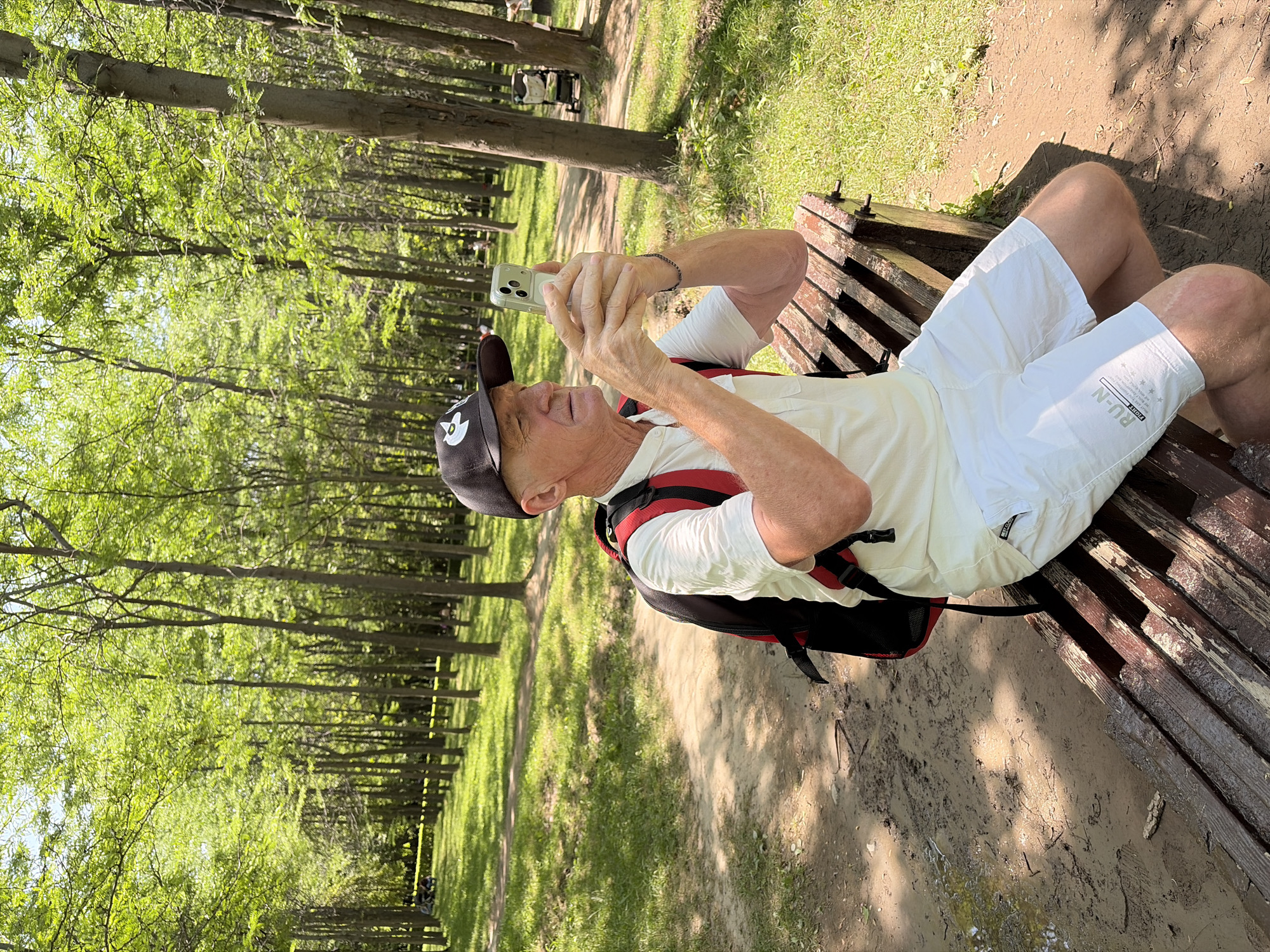 In an outdoor park setting with a dirt path and lush green trees, an elderly man wearing a white polo shirt, white shorts, a black baseball cap with a white logo, and a red and black backpack is sitting on a wooden park bench. He is holding a smartphone in his hands, appearing to take a picture or record a video. The bench is weathered and has visible metal fasteners. The ground around the bench is a mix of dirt, grass, and scattered leaves. In the background, a stroller and a few other people are visible further down the path, among the trees. The sunlight filters through the dense green foliage, casting dappled shadows on the ground.
