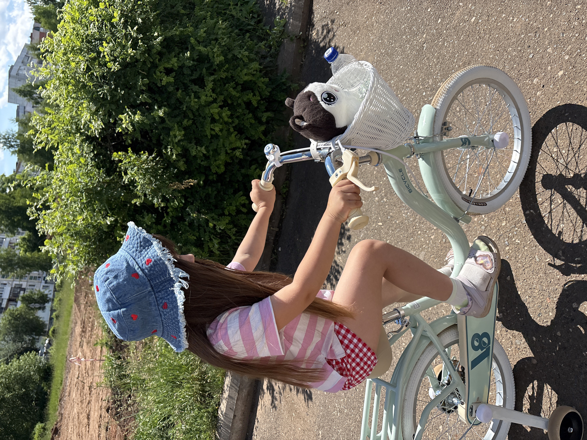 A child is depicted riding a light mint green bicycle on a paved surface under clear, sunny conditions. The child, observed from the right side, wears a denim bucket hat with red heart embroidery and a frayed brim, a pink and white horizontally striped short-sleeved t-shirt, and red and white gingham-patterned shorts. Light-colored sandals are worn over white socks, with the child's right foot positioned on a pedal. The bicycle is equipped with white training wheels visible on the rear left side. A white wicker basket is attached to the handlebars, containing a black and white plush toy with prominent eyes and a clear plastic water bottle with a blue cap. The bicycle frame displays "V8" and "BICYCLE" branding. The immediate environment is an asphalt or similar paved path. The background features dense green foliage, a disturbed earth area on the left, and distant multi-story buildings partially visible through trees under a blue sky with some clouds. Strong shadows are cast, indicative of direct sunlight. The specific location of Chaikovsky, Russia, cannot be determined from the visual evidence presented in the image.