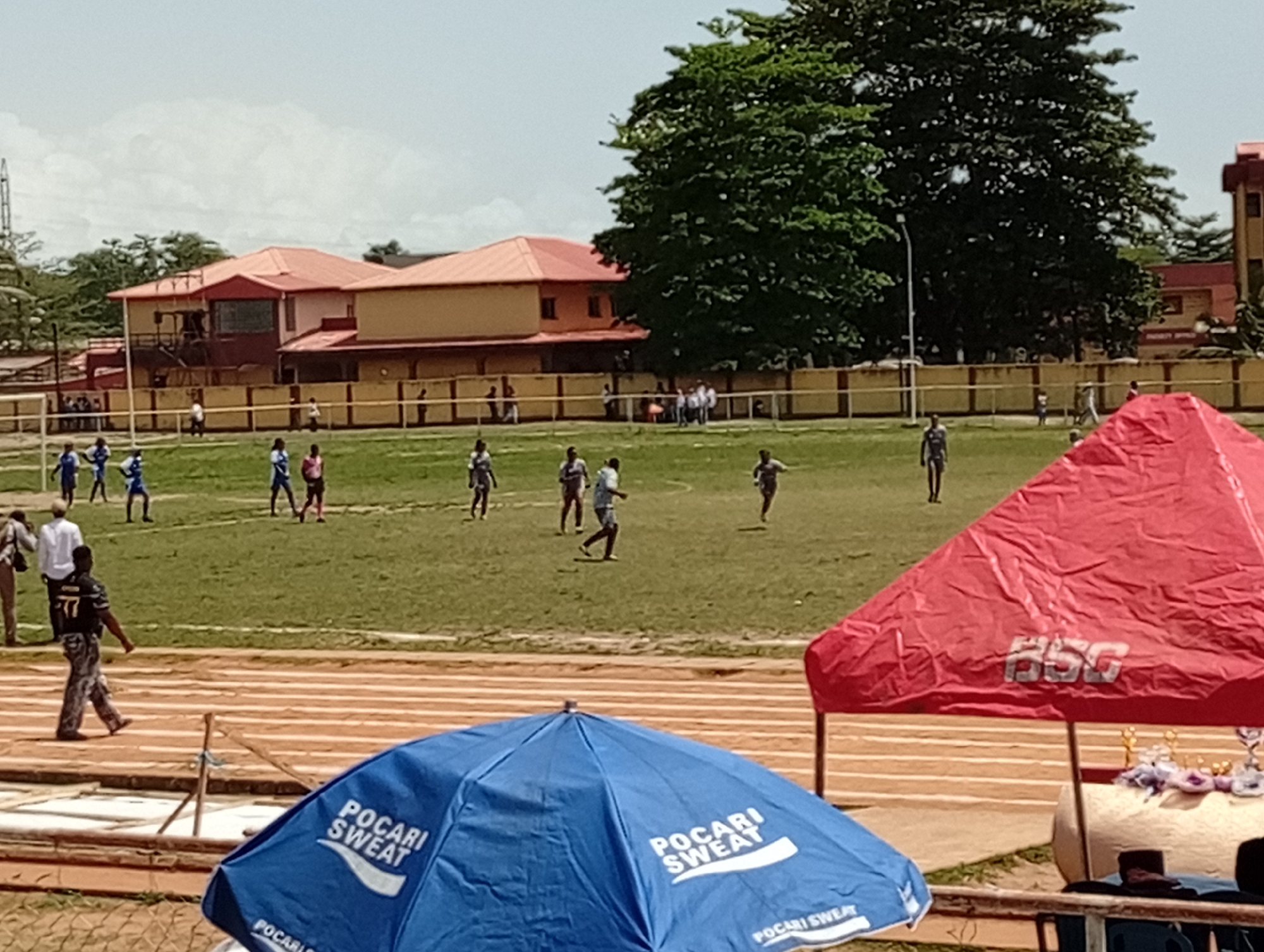 A soccer match is in progress on a grassy field in Unknown, Unknown. The players, clad in athletic gear, are actively engaged in the game. Spectators are visible in the background. A red canopy, labeled "HSG", and a blue umbrella bearing the "POCARI SWEAT" logo are in the foreground. A running track surrounds the field. A building with a red roof and a wall encloses the scene. A large tree is on the right of the image.