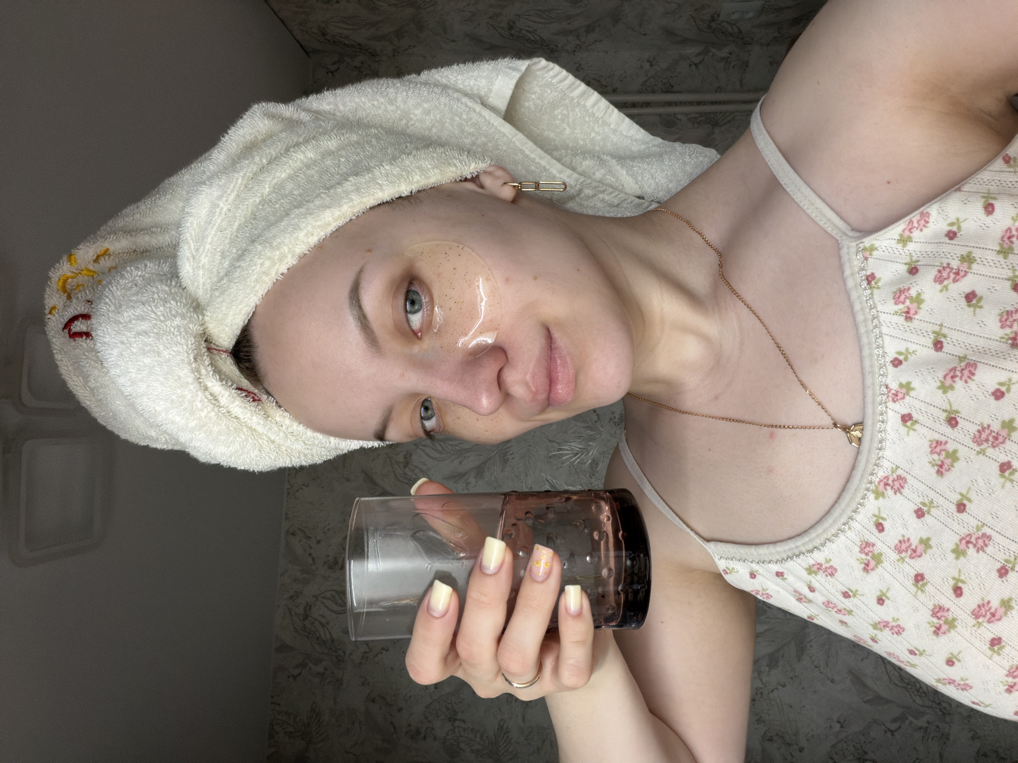 A person is holding a glass with an unknown liquid, indoors. The individual has a towel wrapped around their head, eye masks under their eyes, and is wearing a floral-patterned tank top and a gold necklace. The person also has light yellow nail polish and a ring on their ring finger. The background appears to be a bathroom, with a wall visible behind them. The image does not contain locational information about Reshetnikovo, Russia.
