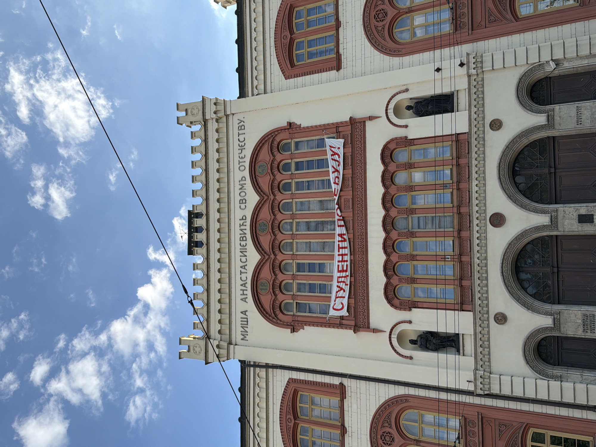 The image depicts the facade of the Rectorate building of the University of Arts in Belgrade, Serbia. The building is predominantly white with decorative red brick accents around the windows and arches. A banner hangs vertically from the upper windows, displaying the word "СТУДЕНТИ" (Students) in red Cyrillic script, followed by "ПО...БУЈУ!" (PO...BUJU!). The building features several arched windows, some with intricate detailing, and two statues in recessed niches. Above the banner, inscription in Cyrillic "МИША АНАСТАСИЈЕВИЋ СВОМ ОТЕЧЕСТВУ" (Misha Anastasijevic to his fatherland) can be read, indicating the building's historical significance. The sky, visible in the upper portion of the image, is blue with scattered white clouds. An overhead power line cuts across the scene.
