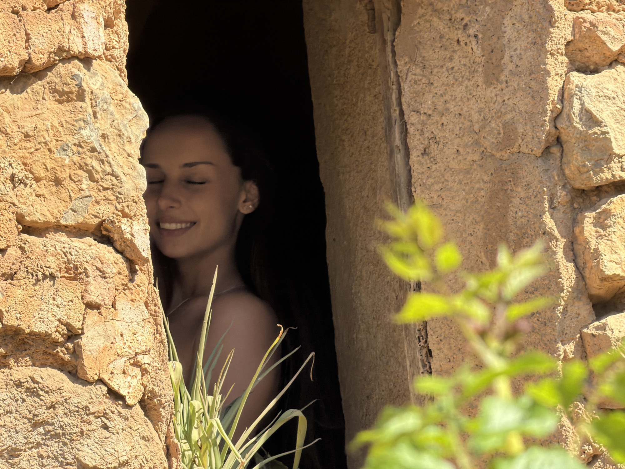 The image depicts a woman with her eyes closed, smiling, positioned within the aperture of a stone structure. She is partially obscured by greenery. The structure appears to be constructed of roughly hewn stone, typical of rural architecture. The surrounding environment is somewhat obscured by shadow, but the presence of foliage suggests an outdoor setting. The image does not provide direct clues regarding its location in Bunyola, Spain, although the stone and flora are consistent with landscapes found in the region.