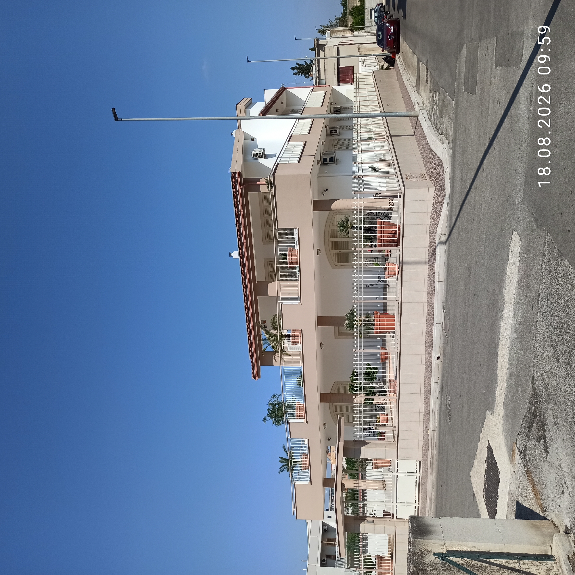 This is a daytime, eye-level, exterior, wide-angle photograph taken on a street in Bitonto, Italy. The main subject is a three-story house with beige and white facade, featuring multiple balconies with white railings and planters. The house has a red-tiled roof and cream-colored shutters. A white metal fence runs along the front of the property. In front of the house, there is a paved street with a pedestrian crossing marked by a white stripe. Several potted plants are visible in front of the house, both on the ground and on the balconies. The sky is clear and blue. In the background, there are other buildings, streetlights, and parked cars, including a red Alfa Romeo. The timestamp "18.08.2026 09:59" is visible in the bottom right corner of the image. There are no people visible in the photograph.