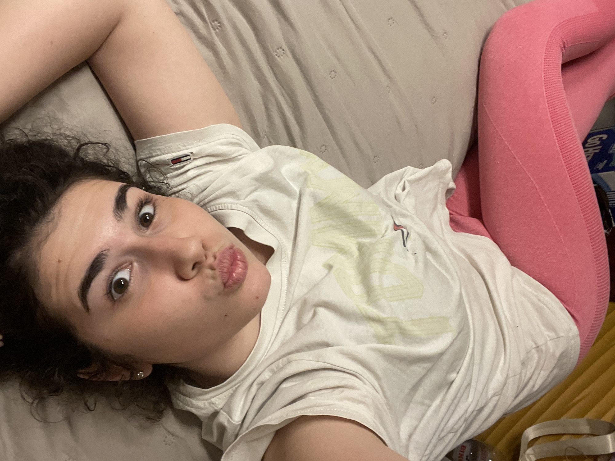 A woman is lying on a bed. She is wearing a white t-shirt and pink leggings. She is making a kissing face. The bed has a beige comforter. A box is located on the floor. The location cannot be confirmed.