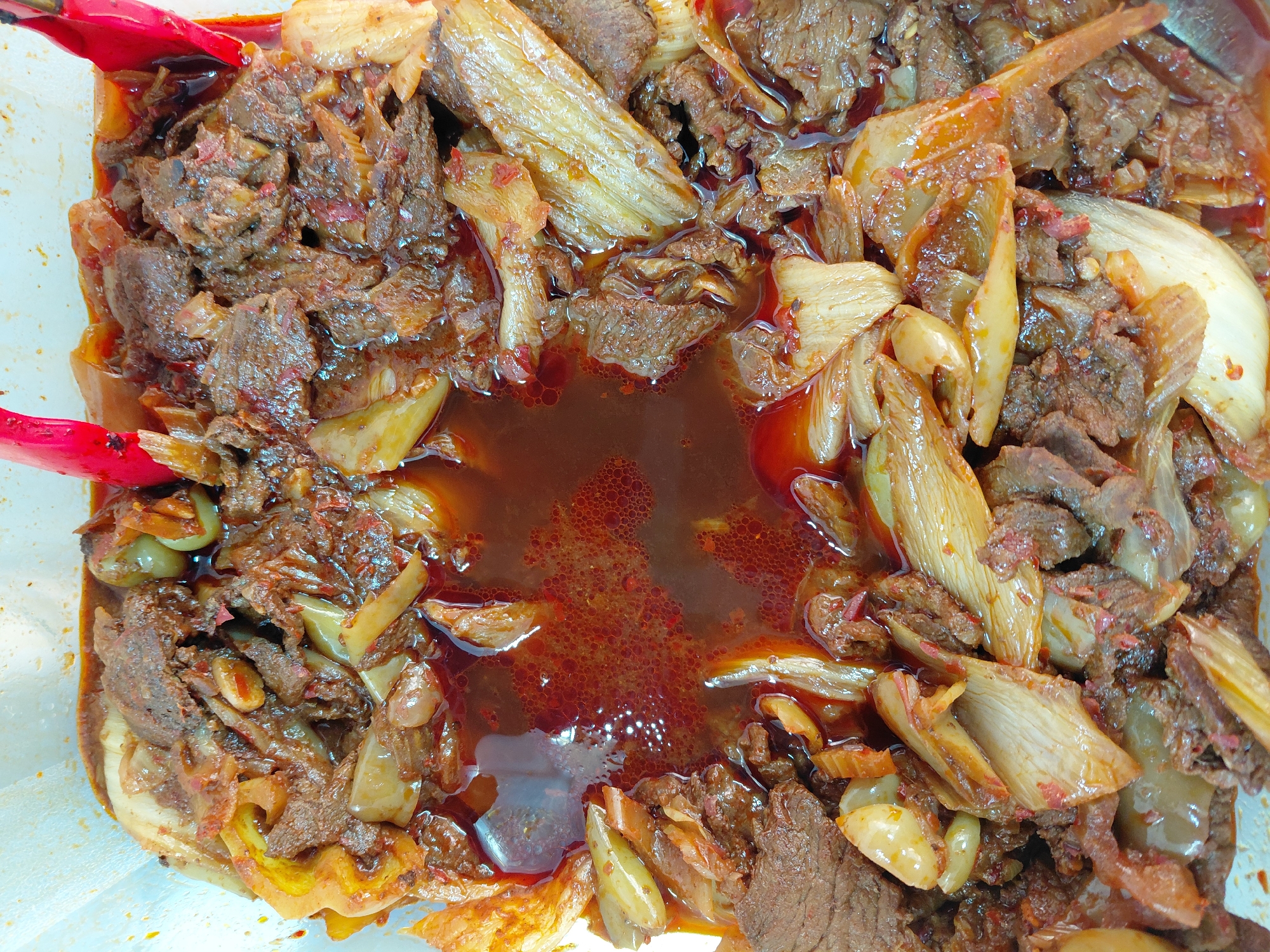 An overhead shot captures a close-up view of a dish of braised beef with onions and what appear to be olives, submerged in a rich, reddish-brown sauce. The food is presented in a white container, and two red-handled tongs are visible at the top edge of the frame, suggesting the dish is ready to be served. The ingredients are a mix of tender, shredded beef, softened slices of onion, and whole green olives. The sauce has a slightly oily sheen with small bubbles visible, indicating it may have been recently cooked. The lighting is bright, highlighting the texture and colors of the food. There is no discernible information in the image to suggest the location within the city of Aktau, Kazakhstan.
