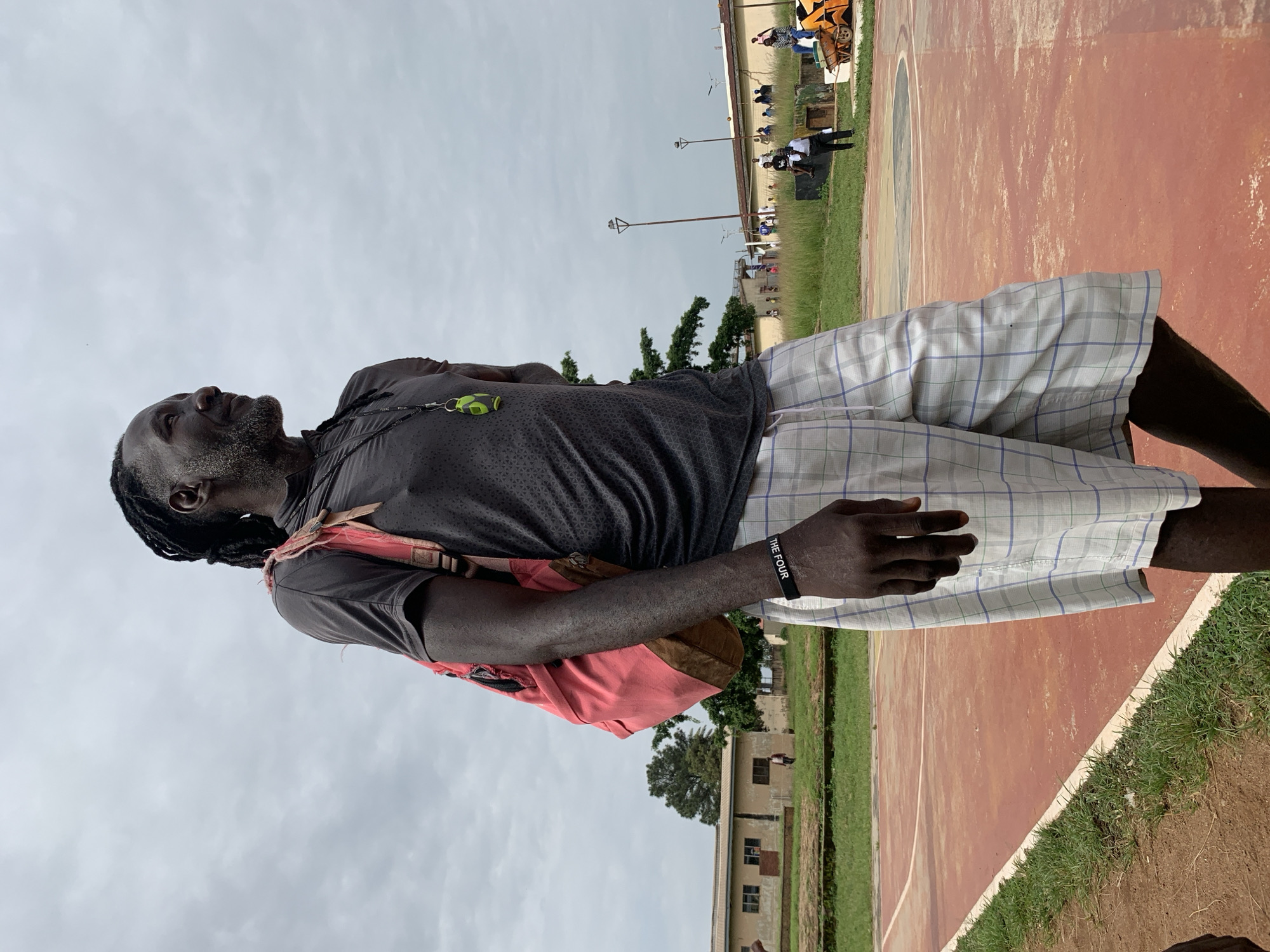 A man with dreadlocks and gray facial hair, wearing a dark gray t-shirt, a pink backpack, and white and blue plaid shorts, stands on a red basketball court. He is looking to the right, with a faint smile. A black wristband with white text that reads "THE FOUR" is visible on his left wrist. A bright green whistle hangs from a lanyard around his neck. The court is surrounded by grass, and in the background, there are buildings, trees, and other people. The sky is overcast. The scene appears to be outdoors in a semi-urban or rural setting. There is no specific indication that this location is in Gura Top, Nigeria, and this information is not present in the visual data.