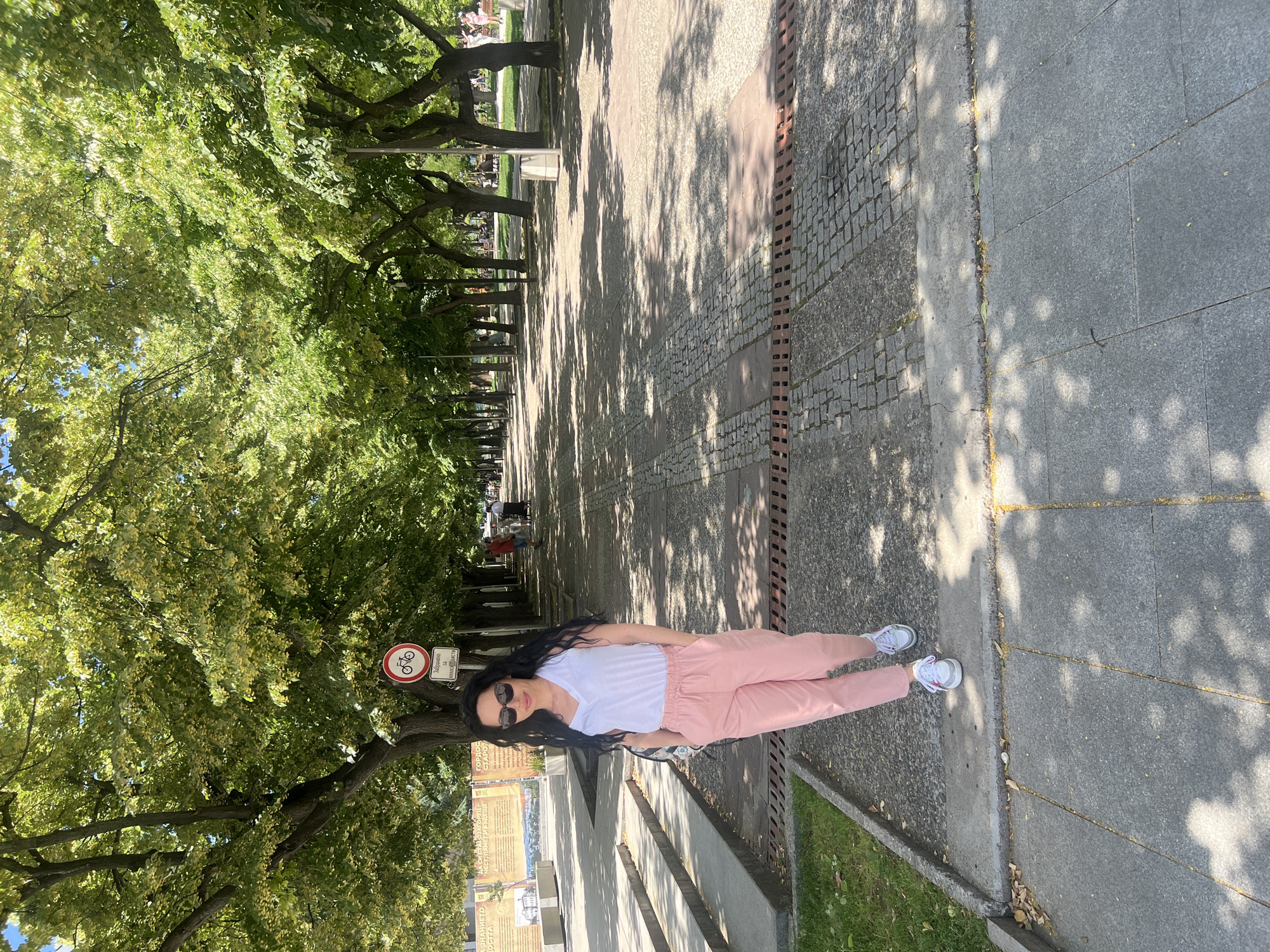A young woman with long dark hair and sunglasses stands on a paved walkway under a canopy of green trees. She is wearing a white V-neck t-shirt and pink pants, with white sneakers. Her hands are in her pockets. Behind her, the walkway extends into the distance, lined with trees and benches, and other pedestrians can be seen. A circular sign with a bicycle symbol and text in Bulgarian is visible on a pole to her left. The area appears to be a park or boulevard, with a grassy area to the left of the walkway and a tiered stone structure. The lighting suggests it is a sunny day, with dappled sunlight filtering through the leaves. This scene is located on the main pedestrian street of Stara Zagora, Bulgaria.