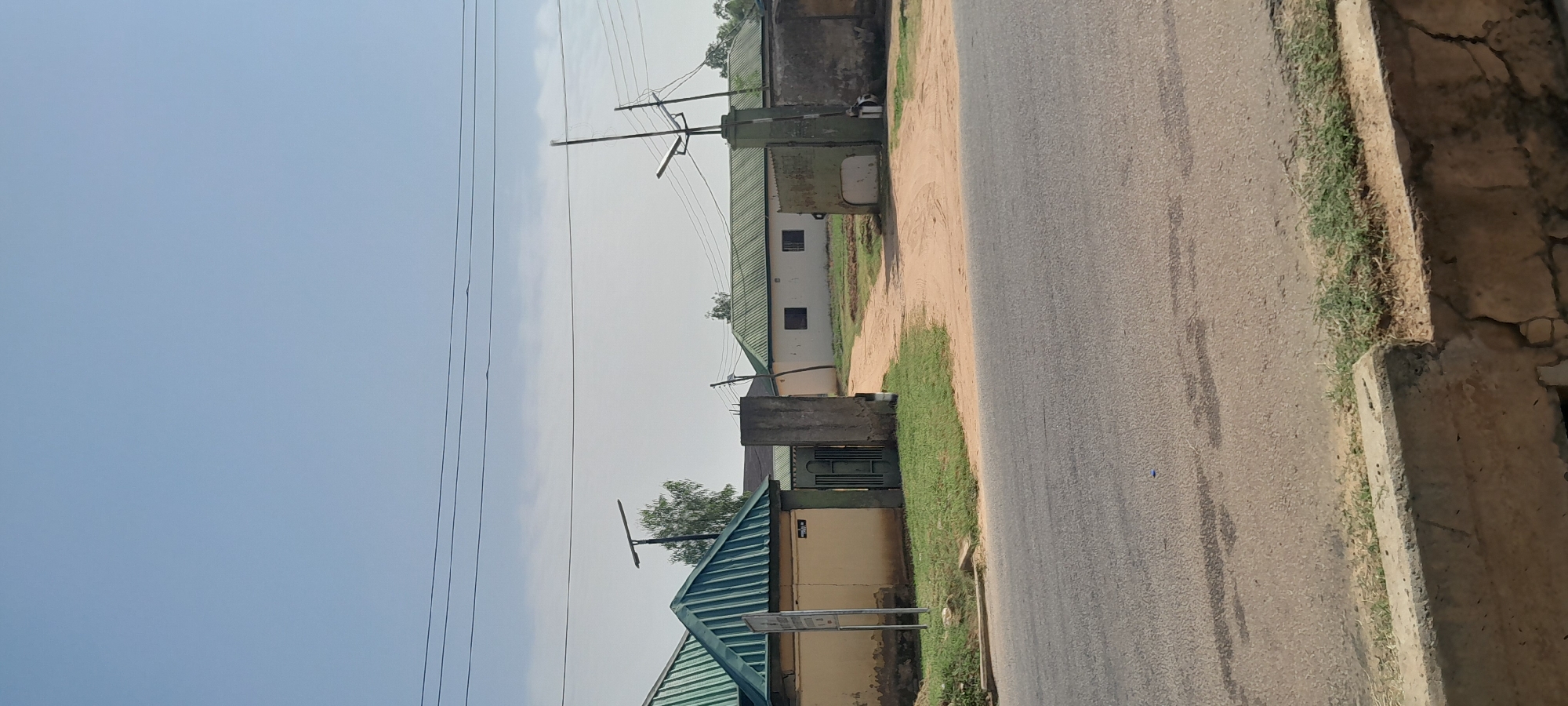 This image depicts the entrance to a residential compound in Jalingo, Nigeria, under a clear blue sky. The compound features a main building with a light-colored facade and a green corrugated metal roof. This building has two dark-colored windows visible. To the left, another structure with a similar green roof is partially visible, featuring a beige-colored wall and a yellow sign. The entrance is marked by a green metal gate, flanked by concrete pillars, one of which appears to be damaged at the base. A dirt path leads into the compound from the road. The road in the foreground is asphalt and shows some wear. Electrical wires and poles are present in the background, suggesting infrastructure in the area. A street lamp is visible in front of the main building. The vegetation consists of sparse grass and a tree. The overall impression is a somewhat modest urban or semi-urban residential setting.