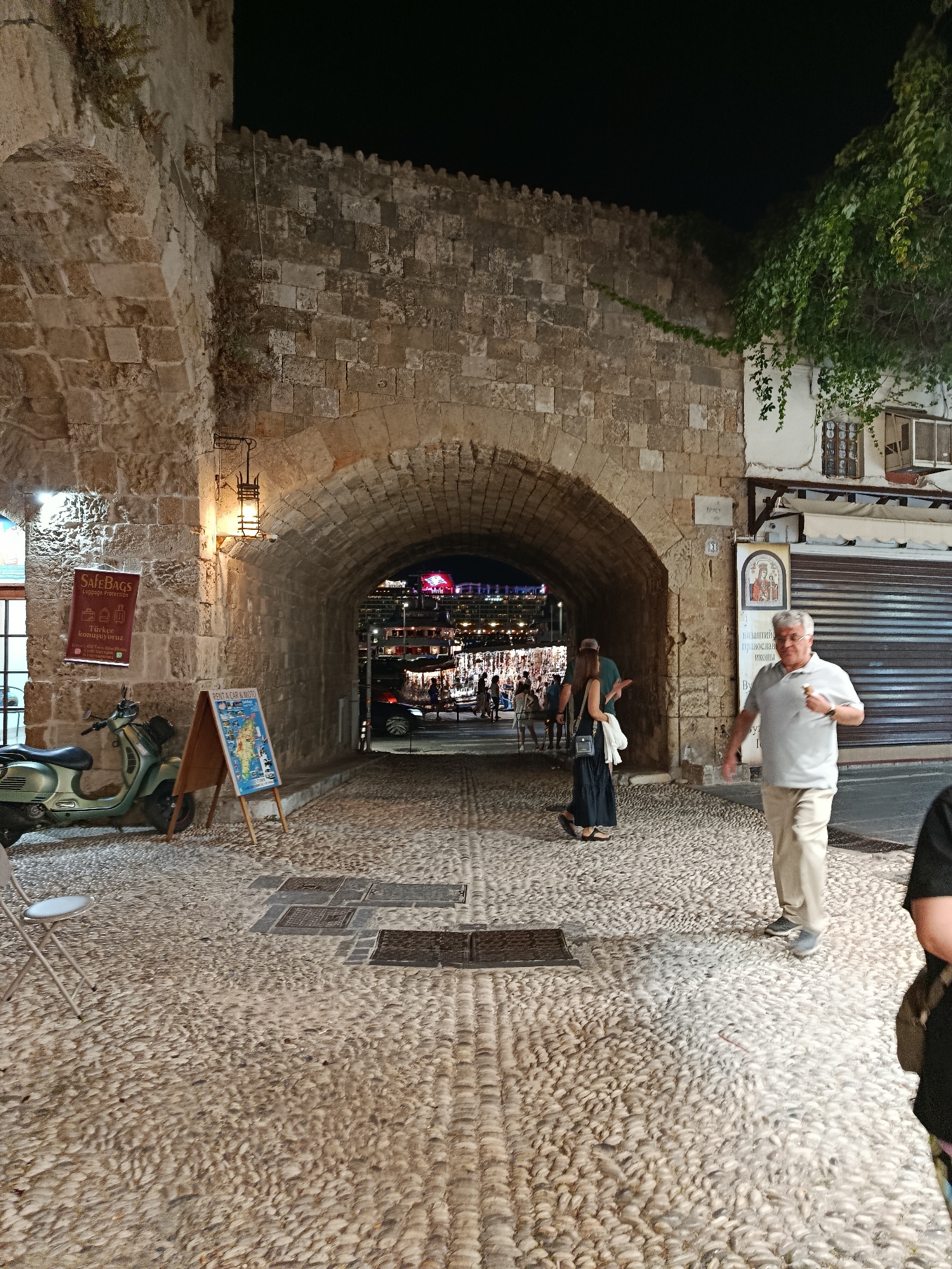 This media file depicts a nighttime scene in a historic European town, likely Rhodes, Greece, given the location context. The image focuses on a cobblestone pathway leading through a large, ancient stone archway.
On the left side of the archway, there is a green scooter parked next to a sign advertising "SAFEBAGS Luggage Protection" and "Türkçe konuşuyoruz," indicating Turkish is spoken. An A-frame sign next to it advertises "RENT A CAR & MOTO" with a map graphic. A single metal chair is also visible on the left.
The archway itself is made of rough-hewn stone, and a vintage lantern hangs from its ceiling, casting a warm glow. Through the arch, a brightly lit area is visible, suggesting a commercial street or square with shops and possibly restaurants, with people milling about.
Several people are visible in the image. An older man in a light grey polo shirt and khaki pants walks towards the viewer on the right side of the pathway, holding something in his hand. Further into the archway, a woman in a long black dress and a man in a green shirt are walking away from the camera. Other figures are indistinct in the background.
The ground is paved with smooth, rounded cobblestones, and there are several metal manhole covers or grates embedded in the path. The overall atmosphere is that of a lively evening in a tourist-oriented, historical location. The architecture suggests significant age, with the stone construction and the style of the archway. The presence of multiple languages on the signs hints at a cosmopolitan tourist destination.