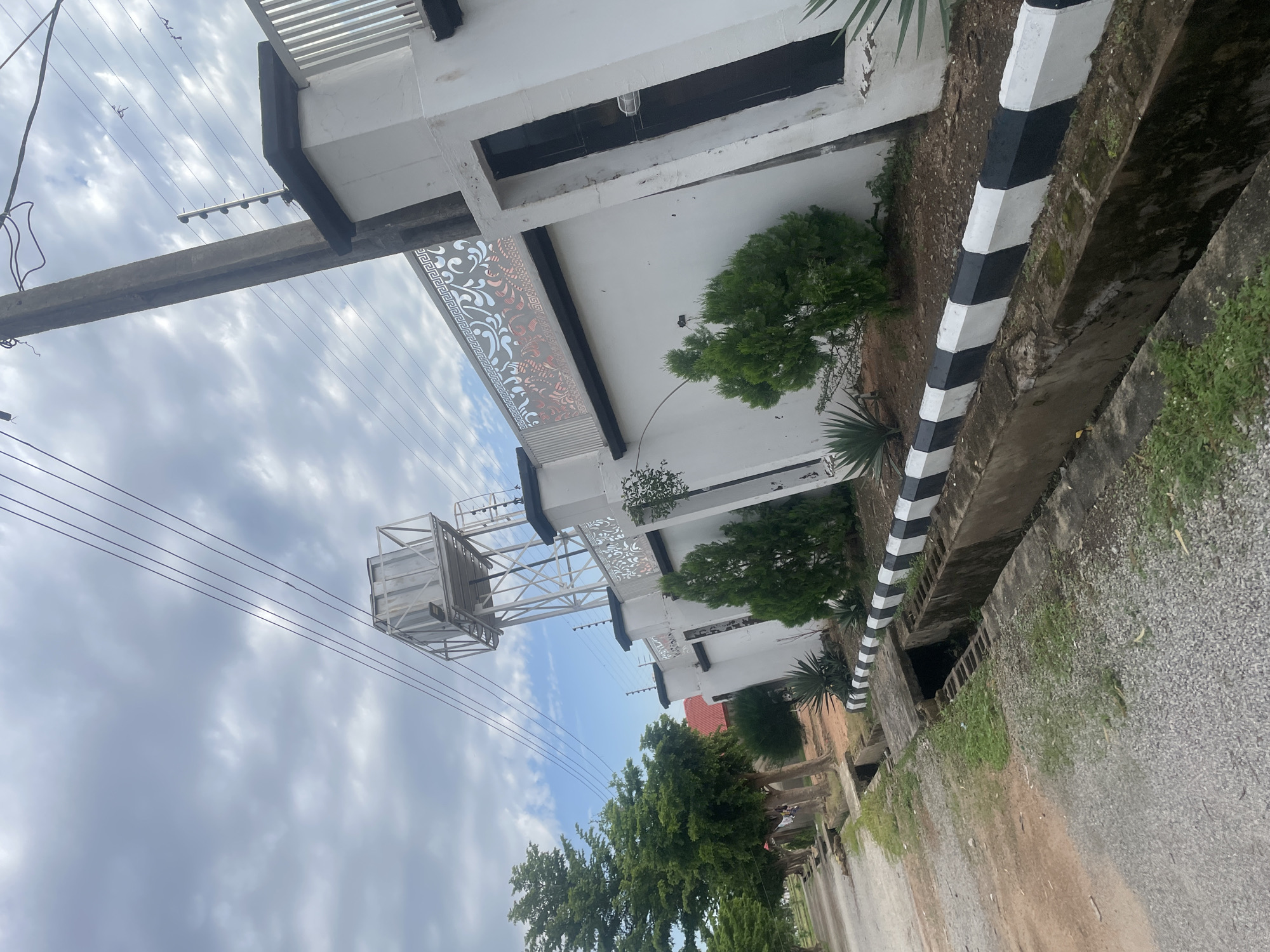 The image depicts a street scene in Jauro Yinu, Nigeria. A white wall with black and white striped coping runs along the right side of the frame. Decorative metal fencing with an intricate pattern is visible along the top of the wall. A concrete utility pole stands to the left of the wall, with multiple electrical wires extending across the sky. Behind the wall, a water tower is visible. The foreground shows a gravel road and a drainage ditch. Several small evergreen trees and some other vegetation are planted along the base of the wall. The sky is cloudy with patches of blue.