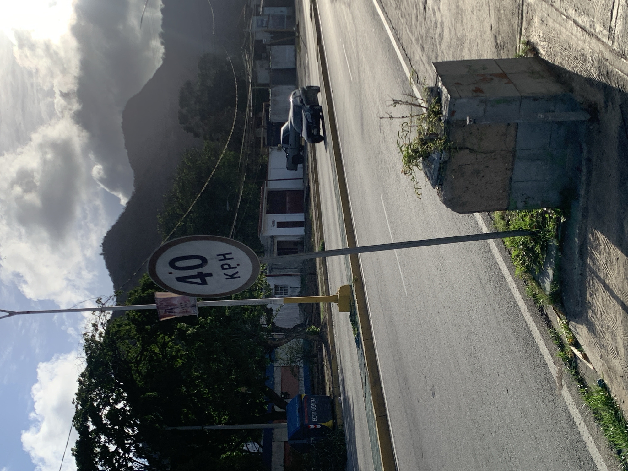 The image depicts a roadside scene in El Valle del Espíritu Santo, Venezuela. In the foreground, a speed limit sign indicating "40 KPH" is prominently displayed on a pole. Adjacent to the sign, on the sidewalk, is a weathered, rectangular concrete planter box with sparse vegetation growing out of it. The road itself appears to be a multi-lane street with yellow and white lane markings. A dark-colored car is in motion on the road, to the right of the speed limit sign. Further in the background, there are several buildings and a blue dumpster labeled "ECOLÓGICA". Trees with dense foliage are visible on the left side of the image. The scene is set against a backdrop of hills or mountains, partially obscured by clouds, under a bright sky. The lighting suggests it is either early morning or late afternoon, with sunlight casting shadows.