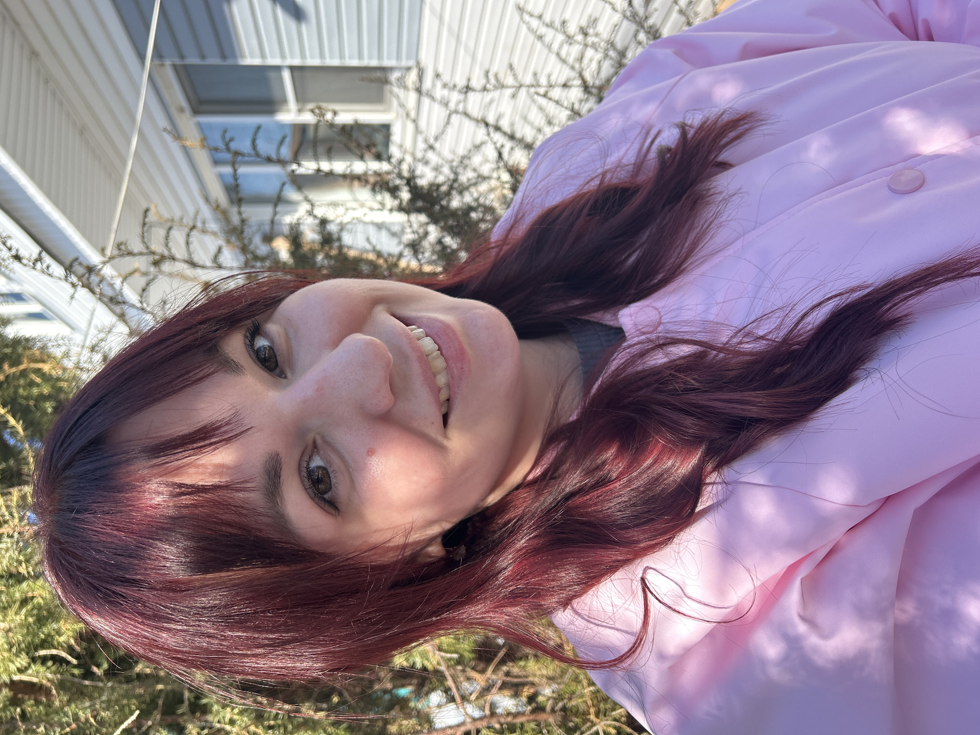 A woman with maroon hair is smiling at the camera. She is wearing a pink jacket. The background includes some greenery and a white building, possibly a house or shed. The location is unknown and cannot be determined from the given image.