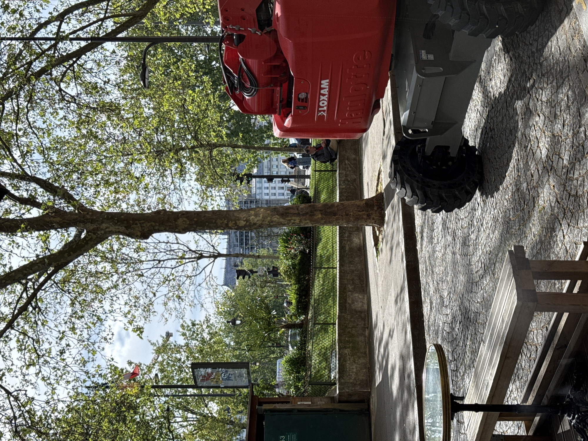 The image depicts a street scene in Paris, France. A cherry picker, branded "LOXAM Haulotte," is parked on the sidewalk. A tree trunk and its branches extend across the foreground. Beyond the sidewalk, a park is visible, with people sitting on benches. Buildings can be seen in the distance. A park map or sign is visible on the left side of the image. A table with a mirror stands next to a bench on the right side.