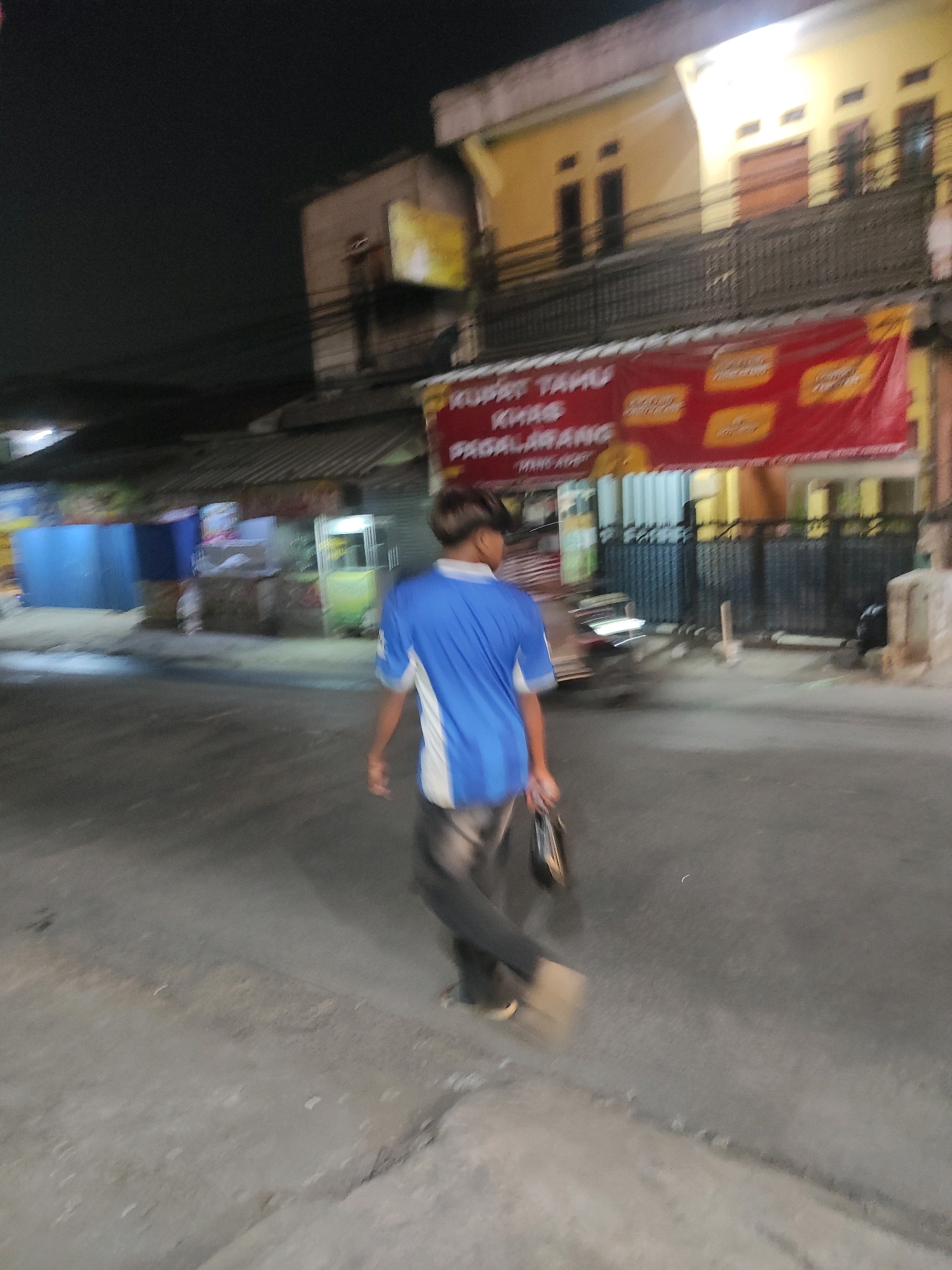 In Padalarang, Indonesia, at night, a person wearing a blue shirt and dark pants walks away from the camera on a paved street. The person is carrying a dark bag in their right hand. In the background, there are buildings with illuminated windows. A red banner hangs across the front of a building, displaying text in yellow and white. Various stalls or shops are visible along the street, some with colorful signs and lights. The overall scene suggests a bustling street at night, possibly a market or commercial area.