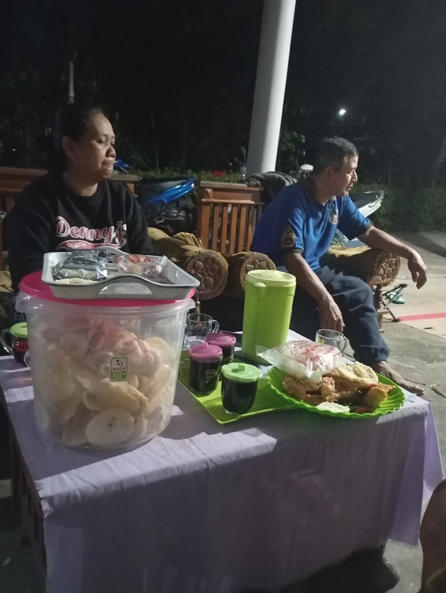 In the city of Wonogondo, Indonesia, a man and a woman are seated at a table outdoors at night. The woman, wearing a black sweatshirt with white lettering, sits to the left, looking to her right. The man, dressed in a blue polo shirt and dark pants, is seated to the right, facing forward and slightly to his left. A white cylindrical object is partially visible to his right.
On the table, a large clear plastic container filled with light-colored snacks, possibly crackers or chips, is prominent. A white tray with various food items, including what appears to be meat and sauces, is placed on top of the container. A green serving tray holds a plate of food with rice, fried items, and some vegetables, along with two small cups containing dark liquid and one cup with a pink lid. A green container with a lid is also on the table. The table is covered with a light purple cloth. Wooden benches are visible in the background, along with parts of what could be a motorcycle. The setting appears to be casual, perhaps a roadside eatery or a gathering at a residence.