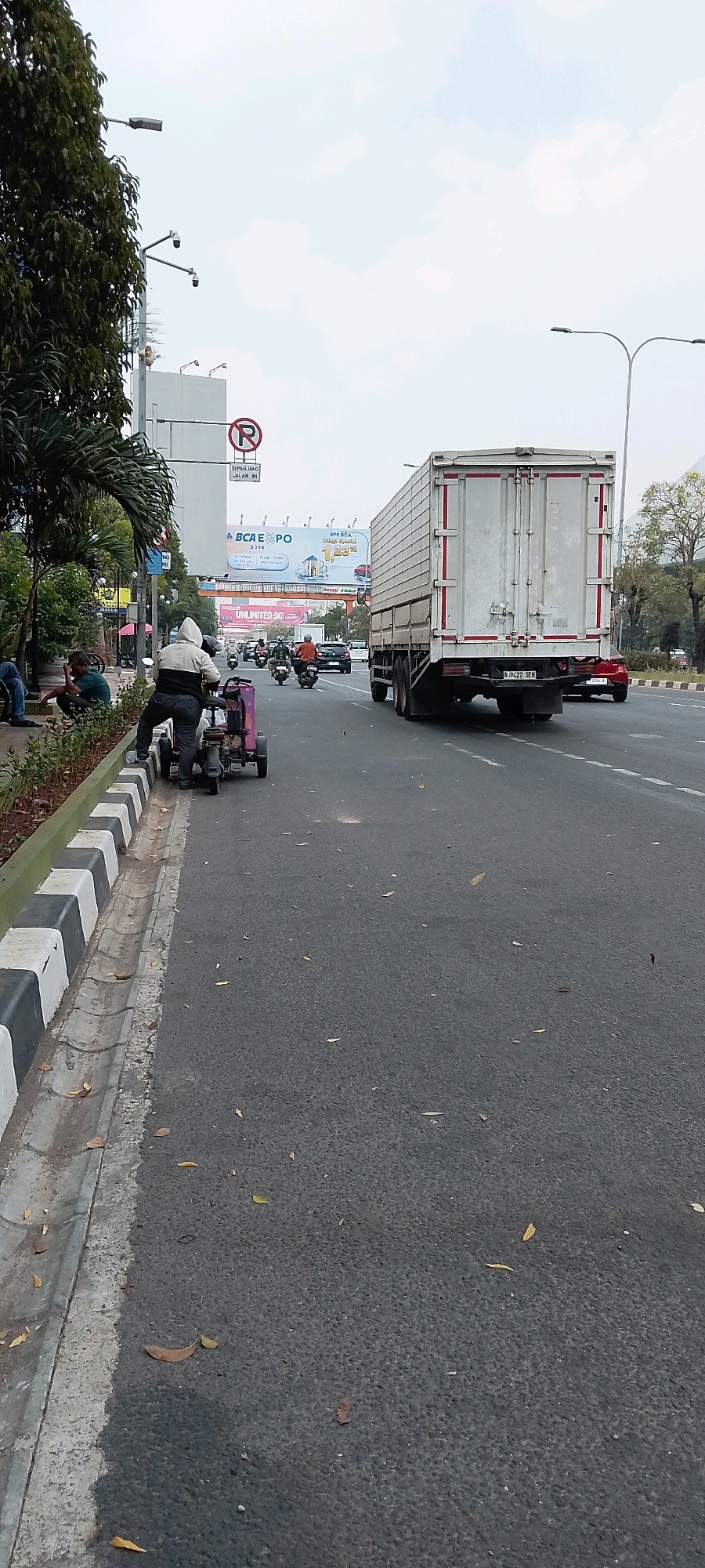 The image depicts a street scene in Bekasi, Indonesia, on an overcast day. In the foreground, a man is standing next to a small, pink three-wheeled vehicle, possibly a modified scooter or a small utility cart, which is parked on the side of the road. The vehicle has a single seat and appears to be manually operated.
To the left of the vehicle, a striped concrete curb marks the edge of the road, with a drainage channel running alongside it. Further to the left, several people are seated on the grass and sidewalk, some appearing to be resting or socializing. Lush green trees and foliage line this area.
The road itself is paved asphalt with white dashed lines indicating lanes. Traffic is present, with several motorcycles and a large white truck visible in the middle ground and background. The truck, a box truck, is moving away from the viewer. A red car is also visible to the right of the truck.
In the background, there are several large billboards and signs. One prominent sign features the logo "BCA EXPO". Other signs are less legible due to distance and resolution, but one appears to advertise "UNLIMITED 5G". Overhead structures, possibly pedestrian bridges or elevated walkways, are also present. Streetlights and traffic signals are visible, consistent with an urban environment. The sky is mostly filled with clouds, indicating a diffused light condition.