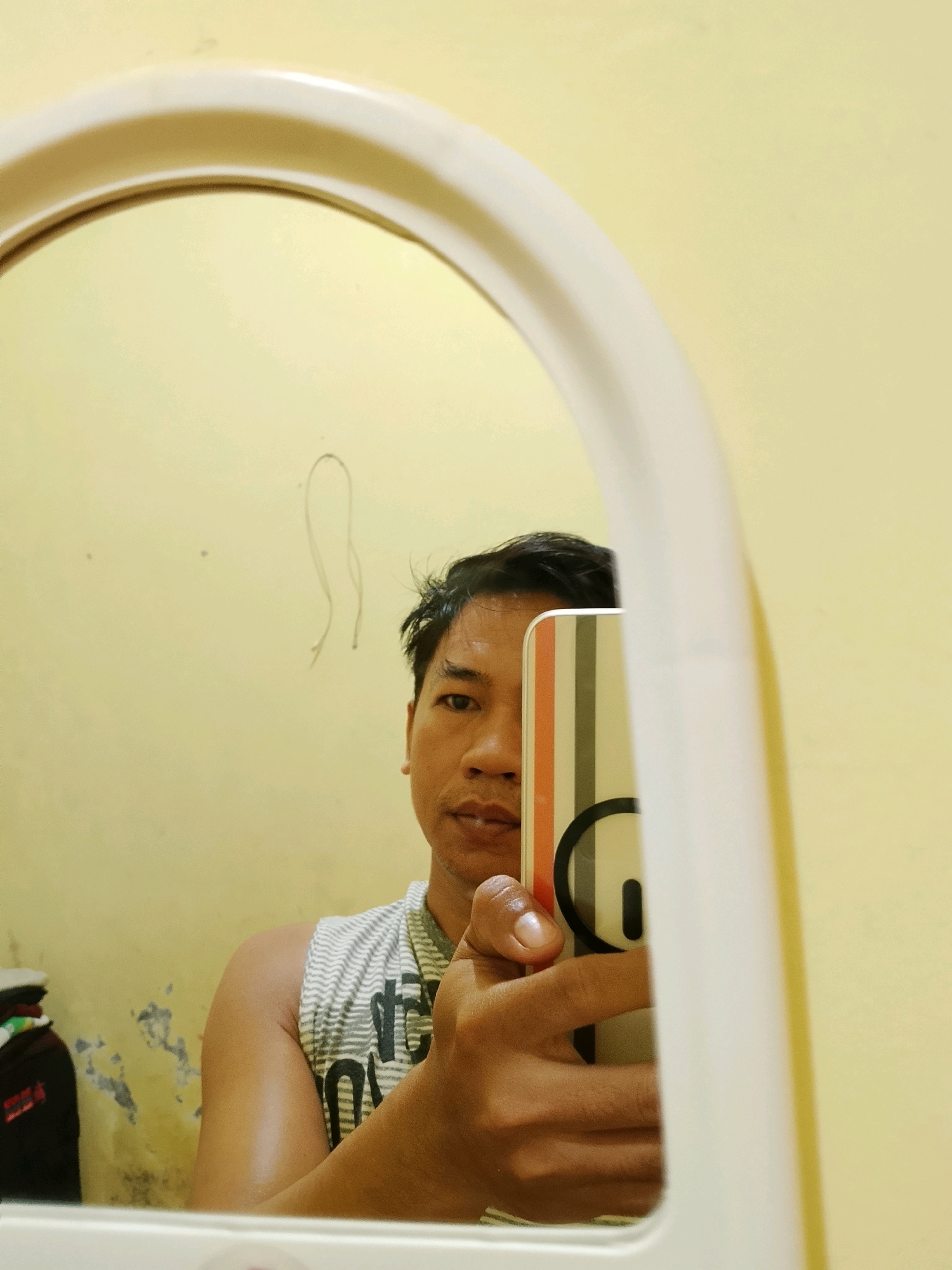 A person is taking a mirror selfie in what appears to be a room with yellow walls. The person is holding a phone with a colorful striped case. The person is wearing a white tank top with black stripes and text. In the reflection, there is a faint drawing on the wall and a stack of clothes on the left. The mirror has a thick white frame. The lighting is soft. The image location is in the city of Pelaihari, Indonesia.