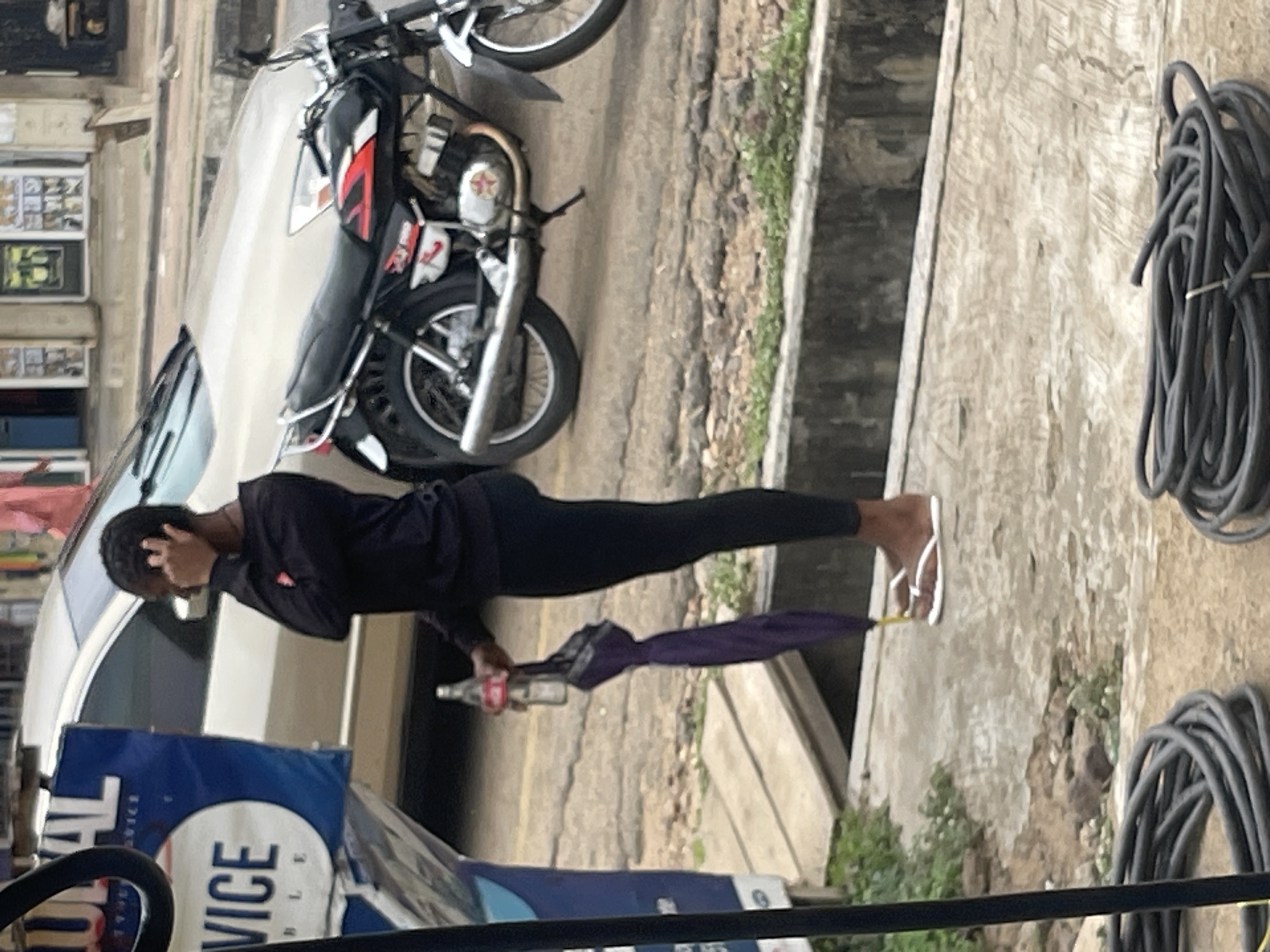 A side profile of a young Black person wearing a black long-sleeved shirt and black leggings stands on a concrete sidewalk holding a bottle of Coca-Cola in their right hand and a closed purple umbrella in their left hand. They are wearing white flip-flops and have their right hand up to their face, covering their ear or scratching their head. Behind them, a tan car is parked, and a black motorcycle is parked to the right of the car. A blue sign with white lettering, partially obscured by a black pole, is to the left of the person. In the foreground, two large coils of black cable lie on the ground. The background shows a building with windows and what appears to be a street and more buildings. The ground is wet, suggesting it may have rained recently.