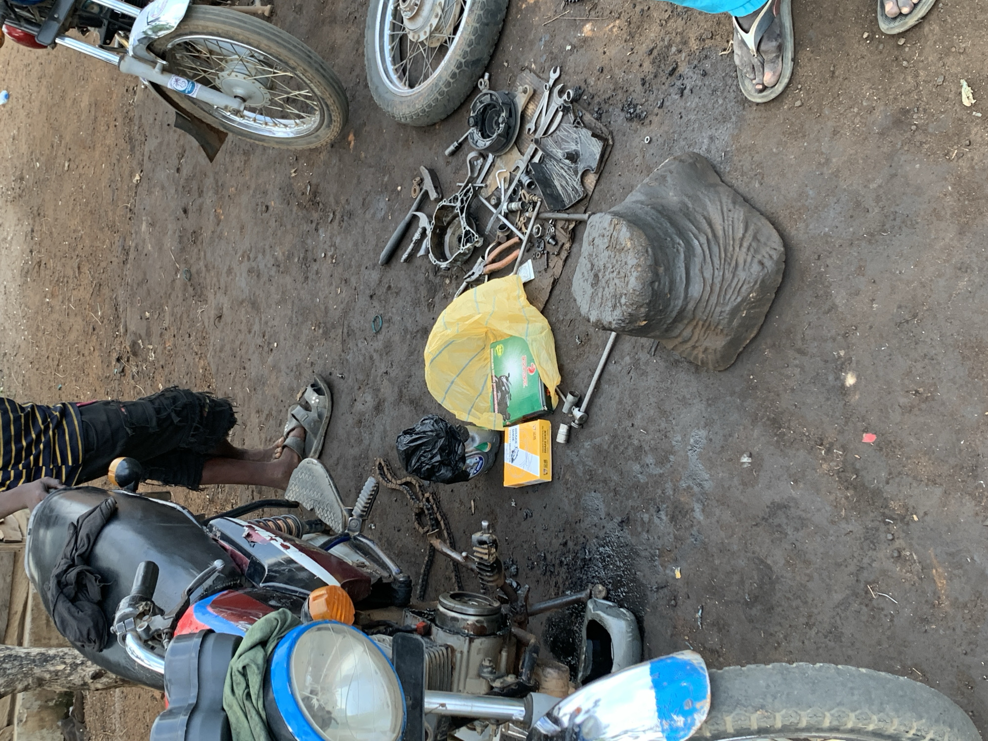 The image captures an outdoor scene at what appears to be a motorcycle repair area in Gbeleko, Nigeria. The ground is unpaved, consisting of packed dirt and some debris.
Multiple motorcycles and their components are the central focus. In the bottom center, a partially disassembled motorcycle is prominent, with its engine exposed, a chain lying next to it, and the front wheel and headlight visible. Another motorcycle's front wheel and fender are seen in the top left corner. A third wheel, possibly detached, rests near the top center.
Several tools are scattered on a dark surface and directly on the ground, including wrenches, hammers, pliers, and various metallic parts, indicating ongoing repair work. Next to these tools are a yellow plastic bag, a black plastic bag, and two cardboard boxes. One box, green and black, has the words "SPARE PARTS" and a logo (possibly depicting a horse) visible. The other, an orange box, partially shows "SPARK PLUG" and "SPARE...". A large, dark, irregularly shaped object, possibly a heavy block or mold, sits on the right.
At least two people are present, evidenced by their feet in sandals. One person is visible in the bottom left, lying or leaning beside the motorcycle, wearing a black and yellow striped garment and black shorts. Another person's feet are visible in the top right.
The scene depicts an active motorcycle repair or maintenance operation, with parts laid out and tools ready for use. The lighting suggests it is daytime, likely under natural light, and there are no indicators of adverse weather.
