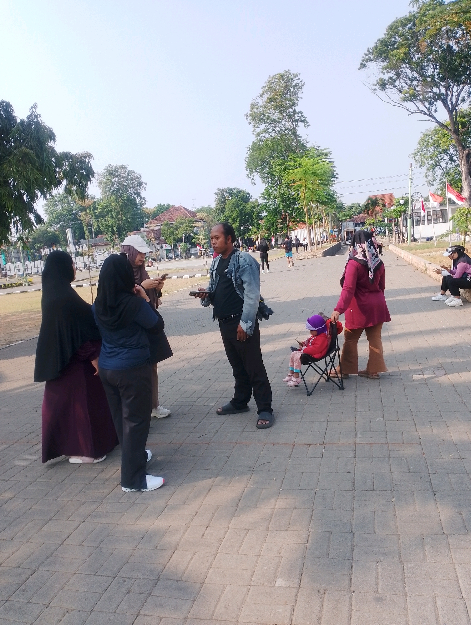 The image depicts an outdoor scene in Probolinggo, Indonesia. In the foreground, on a paved walkway, a man in a denim jacket and black pants is speaking to a group of women. One woman, wearing a dark hijab and a maroon dress, stands to his left. To her left, another woman in a black hijab and dark blue top faces away from the camera. A third woman, wearing a light brown hijab and a cap, stands between them, looking at her phone.
Further into the scene, to the right, a young child sits in a small folding chair wearing a red jacket and purple hat. A woman in a maroon outfit stands next to the child. In the background, there are trees, buildings, and a few people walking. Red and white flags are visible, suggesting a public gathering or event. The environment appears to be a park or recreational area, with a bright, clear sky overhead.