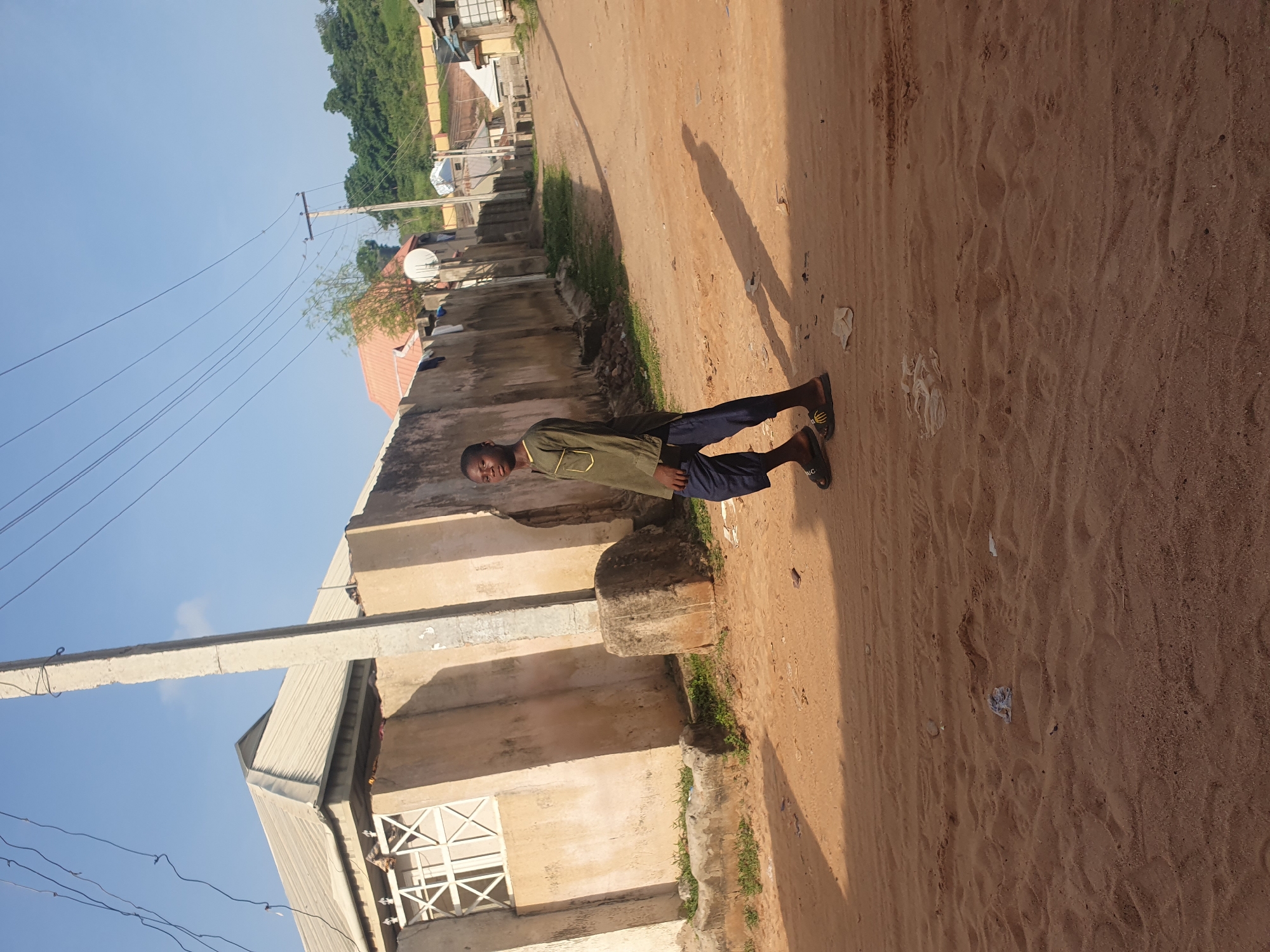 The image depicts a young Black male walking on a dirt road in Lokoja, Nigeria. He is wearing a long-sleeved green shirt with yellow trim and dark blue pants, along with dark sandals. The road is dusty and appears to be unpaved. To the left of the road, there is a beige building with a light-colored, ribbed roof. A white decorative grille is visible on the side of the building. A tall concrete pole with electrical wires runs vertically through the scene. In the background, there are more buildings, trees, and a gentle slope of land. The sky is clear and blue. The sun casts a long shadow of the man onto the road. The overall setting suggests a residential area in a warm climate.