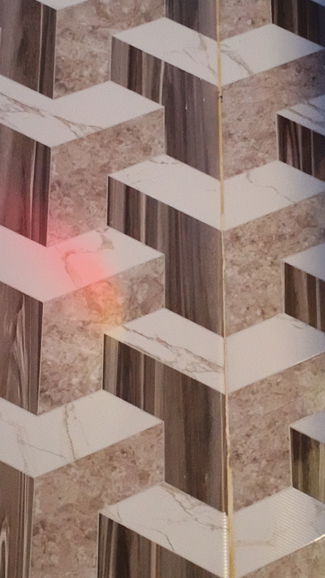 This is a close-up, slightly angled, eye-level shot of a wall with a 3D geometric tile pattern. The tiles are textured, resembling marble and wood grain. The pattern creates a visual effect of stacked and recessed rectangular prisms, giving the illusion of depth. A thin, vertical yellow grout line runs through the center of the image, dividing the pattern. There is a pinkish-orange colored light spot on one of the tiles in the upper left quadrant of the image. The image does not provide any information about the city of Kerep Kidul, Indonesia, or any people, actions, or interactions.