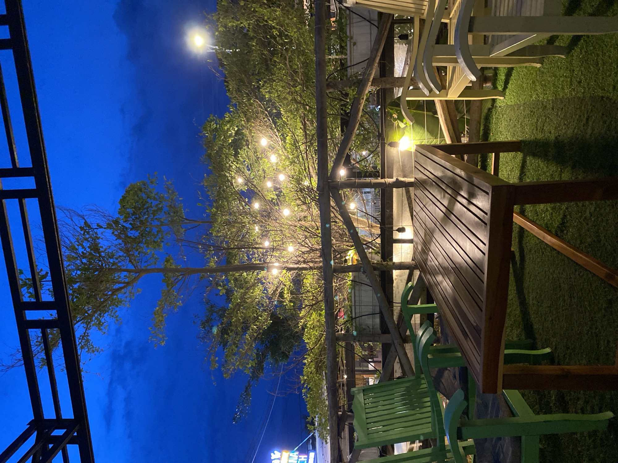 The image depicts an outdoor seating area at dusk, likely a cafe or restaurant patio, located in Phu Thuy Ward, Vietnam. A dark blue sky with scattered clouds forms the backdrop, illuminated by string lights draped across a tree and a distant street lamp. In the foreground, a wooden table is centrally placed on a patch of artificial turf. To the left, two green wooden chairs are positioned, while to the right, two light-colored wooden chairs are visible. A wooden railing separates the seating area from the background, where lush green foliage and more trees are present. A neon sign, partially visible in the upper left corner, suggests the establishment is in a populated area. The overall atmosphere is relaxed and inviting, characteristic of an evening dining or gathering space.