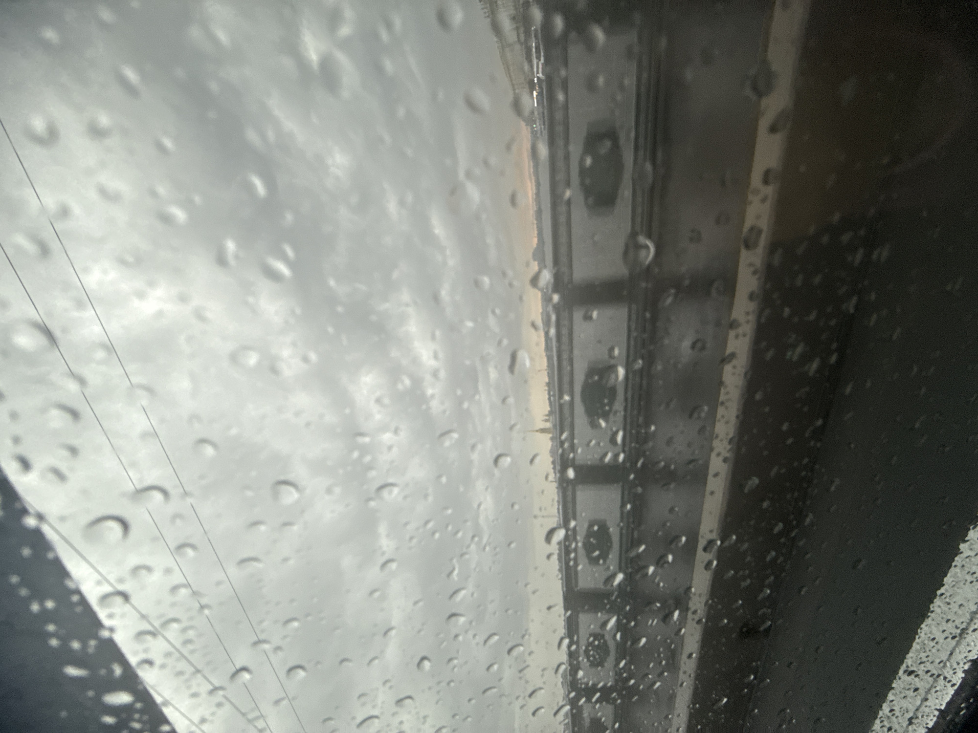 The image shows a view through a rain-streaked window, looking out at a cloudy sky, a bridge, and a city skyline in the distance. The bridge is lined with white balustrades and decorative elements. In the background, a spire, possibly the Admiralty Spire, is visible, suggesting the location is Saint Petersburg, Russia. The overall mood is somber and atmospheric, consistent with a rainy day.