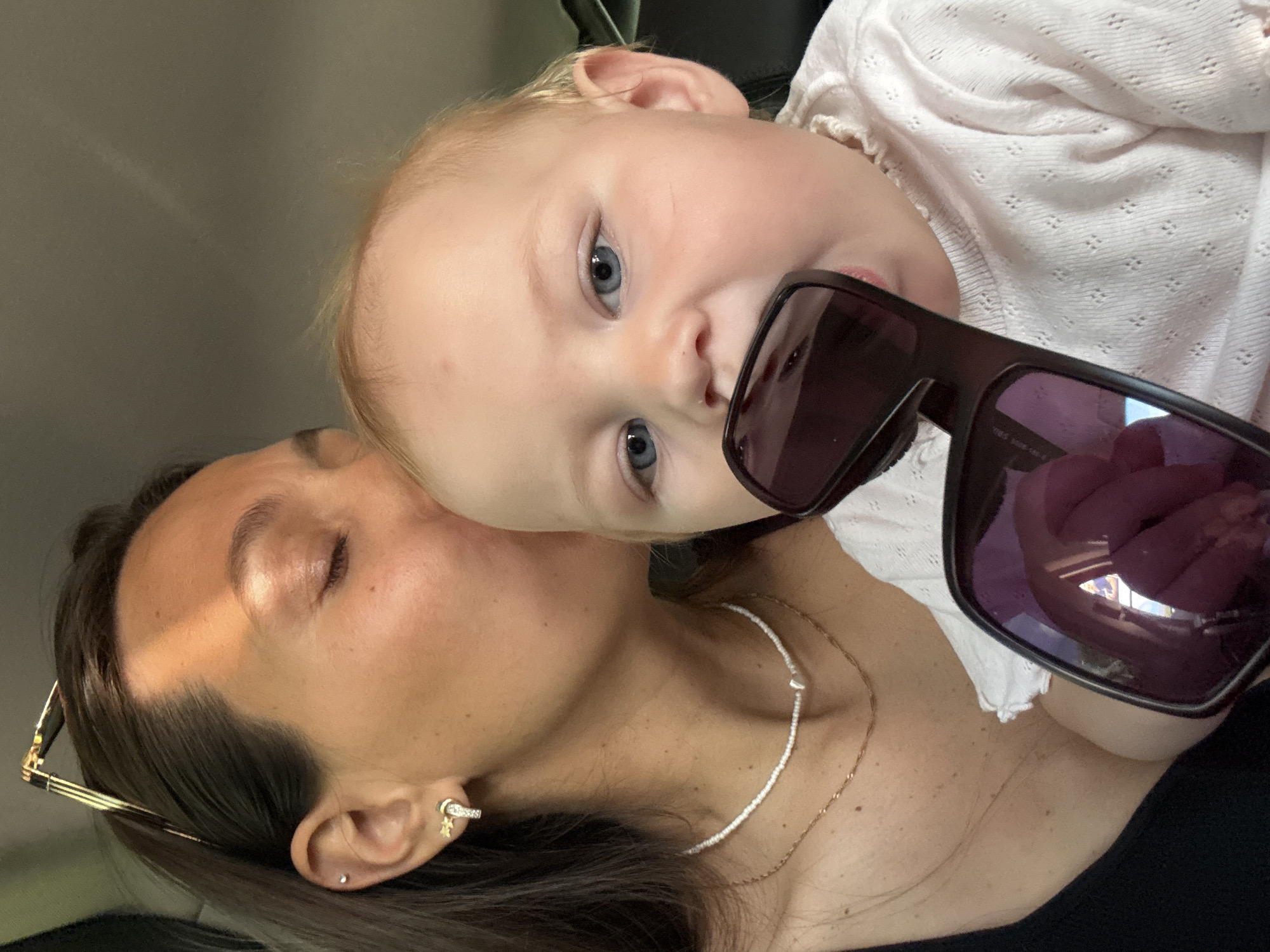 The image depicts a close-up, selfie-style shot of a woman and a baby. The woman, with her eyes closed and her forehead touching the baby's head, is leaning in to kiss the baby. She has dark hair pulled back and is wearing a black tank top, a beaded necklace with a heart pendant, and earrings. The baby, with blue eyes and blonde hair, is looking at the camera and has a pair of sunglasses in its mouth. The baby is wearing a light pink, textured long-sleeved shirt. The background is blurred, suggesting an indoor or car setting. The lighting suggests it is daytime. The image is taken at an unspecified location in the city of Chaikovsky, Russia.