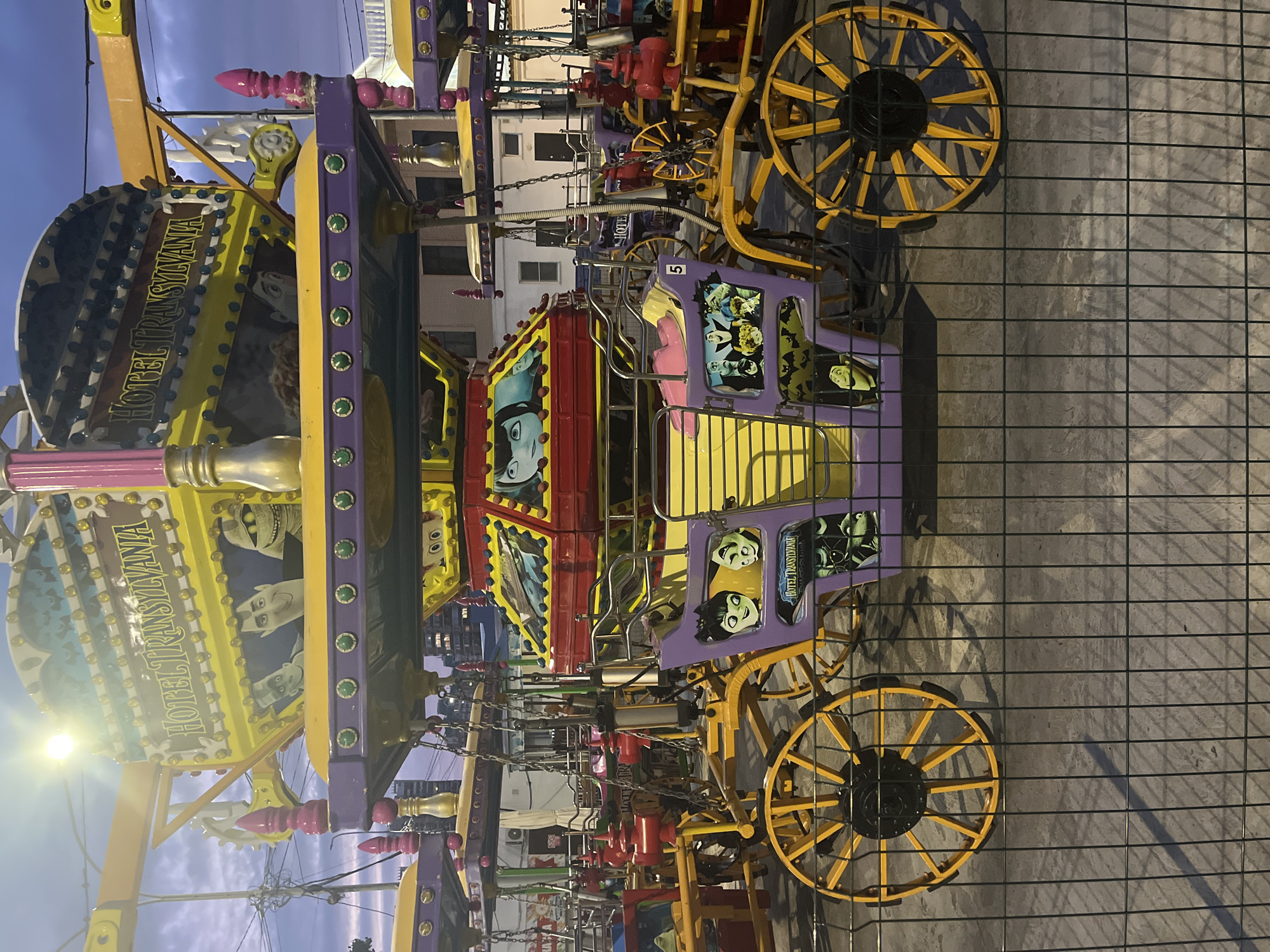 The image shows a carousel ride themed after the movie "Hotel Transylvania" in Krasnodar, Russia. The ride is designed to look like a castle with various carriages. The carriages are purple and yellow and decorated with characters from the movie. The carousel is illuminated by bright lights. The sky is dark blue, indicating it is either dusk or night. The ride is enclosed by a wire fence. There are no people visible in the image.