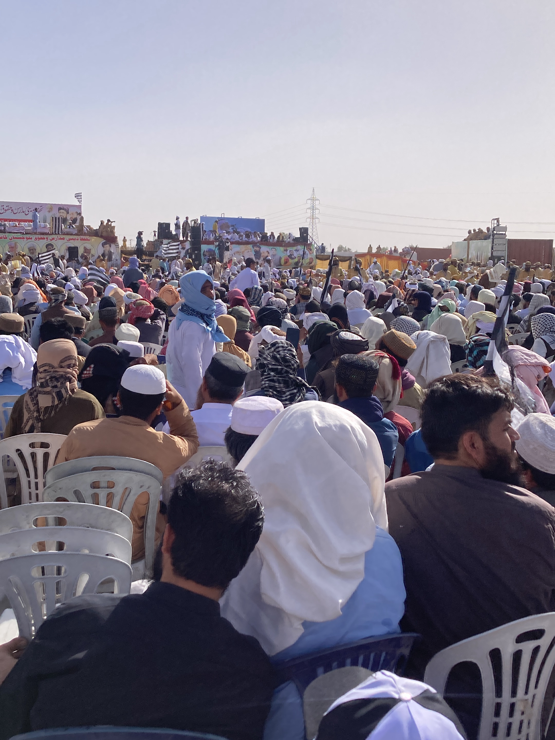 The image depicts a large outdoor gathering or political rally taking place in Pishin, Pakistan, during bright daylight hours under a clear, sunny sky. A substantial crowd, predominantly composed of men, is visible across the scene, filling the foreground, middle ground, and extending towards the background. Many attendees are seated on white plastic chairs, facing a central stage or platform.
The participants are dressed in traditional attire, including shalwar kameez, with a significant number wearing white skullcaps (kufi) or other forms of head coverings and turbans. Some individuals in the crowd also have their faces partially covered with scarves or balaclava-like garments. In the immediate foreground, the backs of several individuals are seen; one person wears a white head covering and a light blue garment, while another has a dark shirt and a beard. A distinctive figure in the middle ground stands out with a light blue head covering and white clothing.
The focal point of the event is a raised stage or platform in the background where several people, likely speakers or organizers, are gathered. The stage is decorated with banners displaying text in Urdu script, though the specifics are hard to decipher from a distance. Some banners and flags visible among the crowd and on the stage feature black and white striped patterns, which are characteristic symbols of certain political or religious parties, such as the Jamiat Ulema-e-Islam (F) (JUI-F). In the far background, an electrical transmission tower stands tall against the horizon, with what appear to be distant structures or buildings. The overall scene conveys a well-attended public event, likely a political or religious congregation.