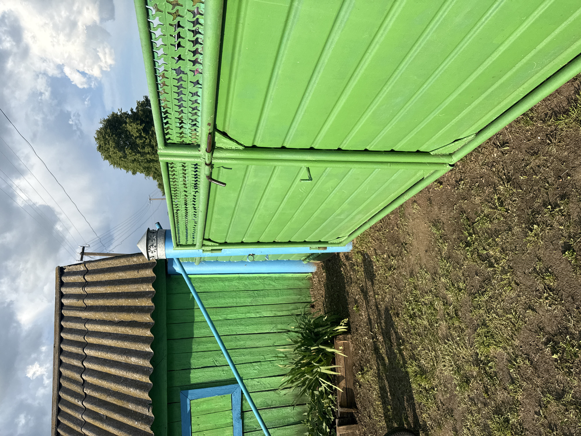 A bright green metal gate with a star-shaped pattern on its upper section stands to the right of the image. The gate is partially open, revealing the interior of a yard. To the left of the gate, a green wooden building with a corrugated roof is visible. A blue window frame is present on the building's wall. In front of the building, a wooden planter box holds lush green plants. The ground is a mix of dirt and sparse grass, with shadows cast by the objects in the scene. The sky above is partly cloudy with dramatic clouds. The scene appears to be in an outdoor rural or semi-rural setting. The location is identified as Чуй-Атасево, Russia.