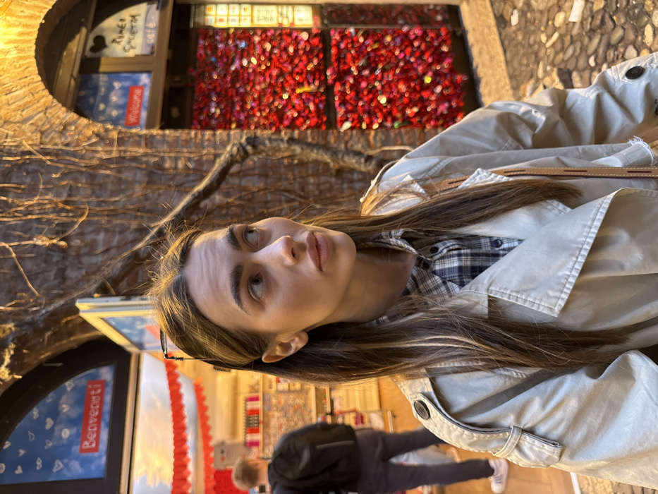 The image captures an outdoor street scene in Verona, Italy, specifically in the vicinity of Juliet's House (Casa di Giulietta). In the foreground, a female subject with long, highlighted brown hair is depicted in a three-quarter profile, looking towards the right side of the frame. She wears a beige trench coat over a blue and white plaid shirt, and eyeglasses are perched on top of her head. To the far left in the mid-ground, a male subject with a black backpack is partially visible from behind, standing within a brightly lit commercial space, likely a shop, marked by a sign displaying "Benvenuti" (Welcome) in Italian and heart graphics. To the right of the female subject, an old brick archway frames a wall densely covered with numerous small, predominantly red, heart-shaped objects or notes. Above this archway, another sign indicates "Welcome" in English and partially visible Italian text, possibly associated with a cafe or landmark. Dark, gnarled vines cling to the brick wall. The ground is paved with cobblestones. The scene is illuminated by a mix of natural and artificial light, creating a warm ambiance.