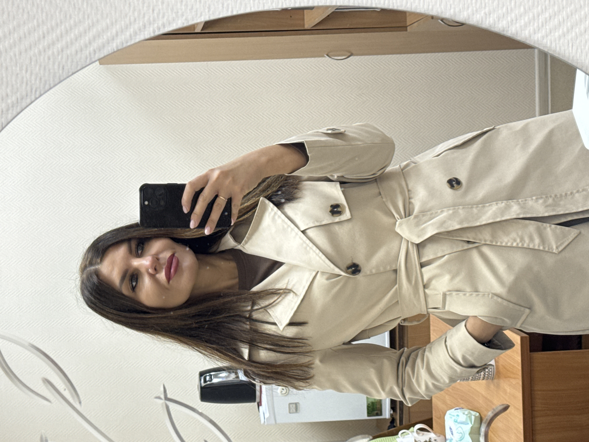 A woman is taking a selfie in a mirror. She has long, dark brown hair, fair skin, and red lipstick. She is wearing a tan trench coat and holding a black smartphone. The background appears to be an indoor setting, possibly a room in a home, with white patterned wallpaper and light wood furniture. Objects visible in the background include a white refrigerator, a black desk organizer, and various other items on a table. The photograph seems to be taken in a residential setting, though the exact location in the city of Chaikovsky, Russia cannot be definitively determined from the visual information provided.