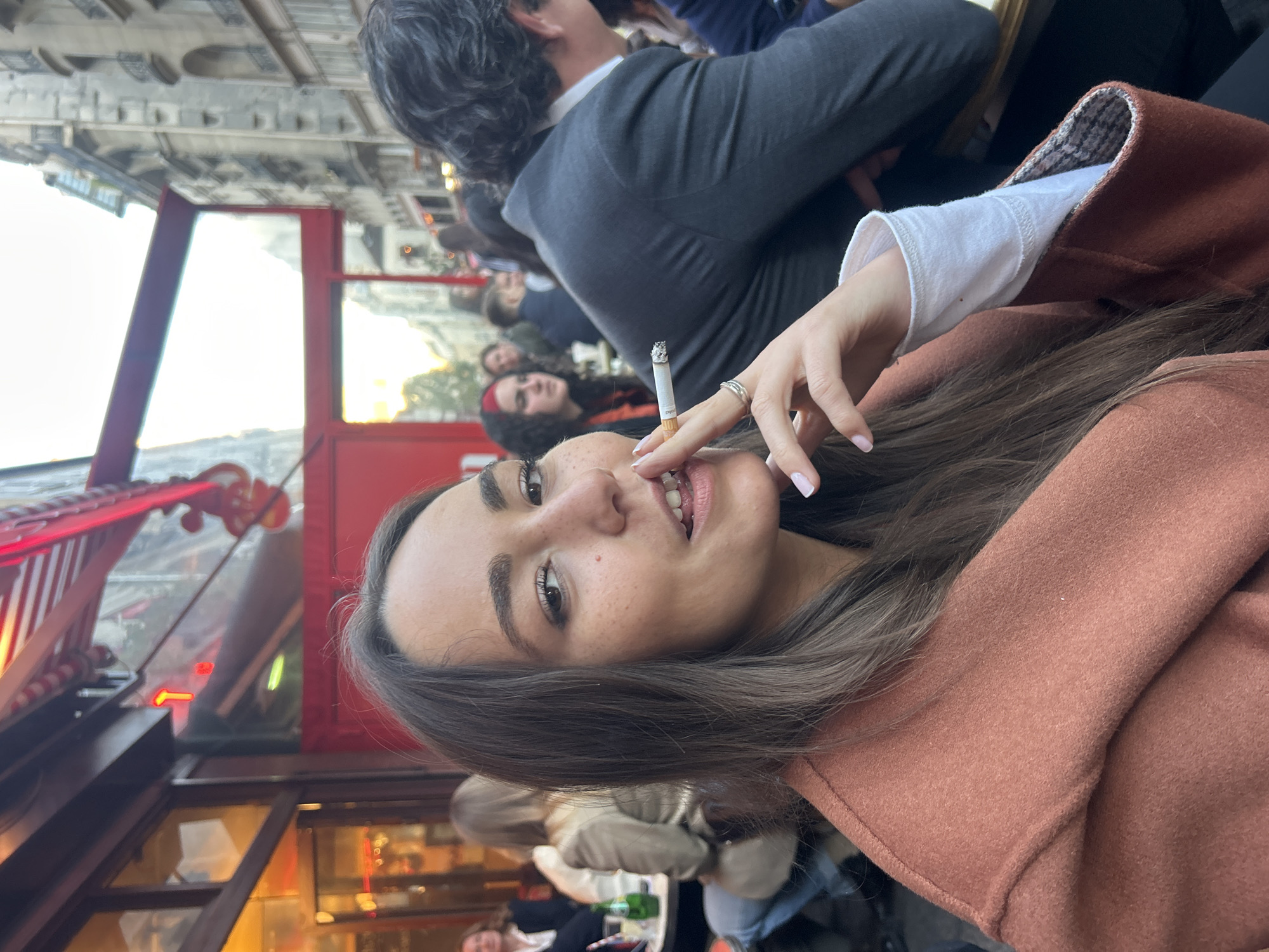 The image depicts a woman in an outdoor setting, holding a lit cigarette to her lips. She has brown hair and freckles, and she is wearing a terracotta-colored coat over a white shirt. A silver ring is visible on her ring finger. In the background, other people are seated at tables, suggesting a cafe or restaurant setting. Red elements of a structure, potentially part of an awning or building facade, are visible. The setting is outdoors, implying a public space.